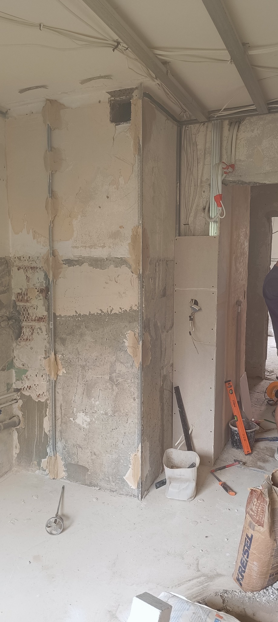 Wnętrze w trakcie remontu. Widoczne ściany z fragmentami starej farby, częściowo pokryte gładzią, oraz zamontowana płyta karton-gips. Przewody elektryczne i narzędzia.