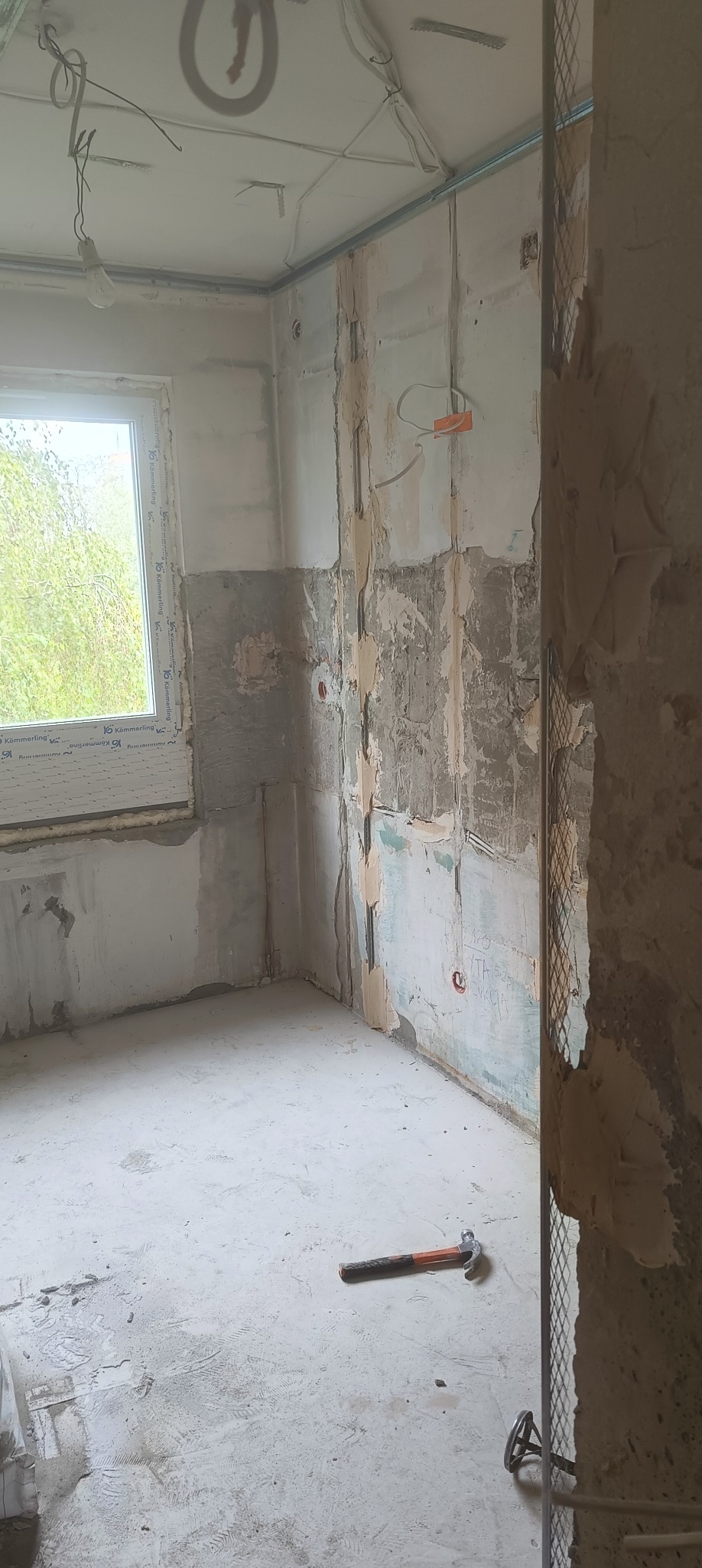 Wnętrze pokoju w trakcie remontu: przygotowane ściany z fragmentami starej farby, widoczne instalacje, siatka wzmacniająca, młotek na podłodze. Okno z widokiem na zieleń.
