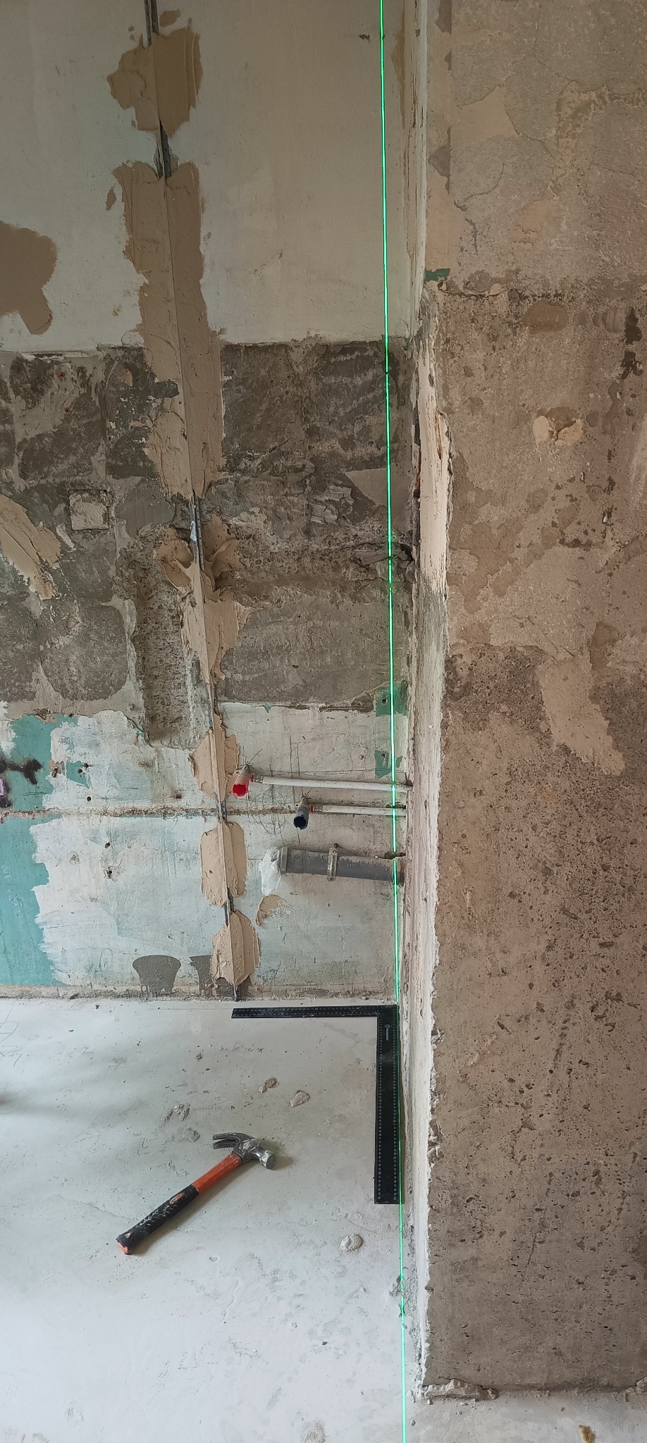 Surowy stan ścian z zielonym laserem pionującym, widoczne ślady po instalacjach, częściowo nałożona gładź, młotek i poziomica na podłodze. Przygotowanie do remontu.