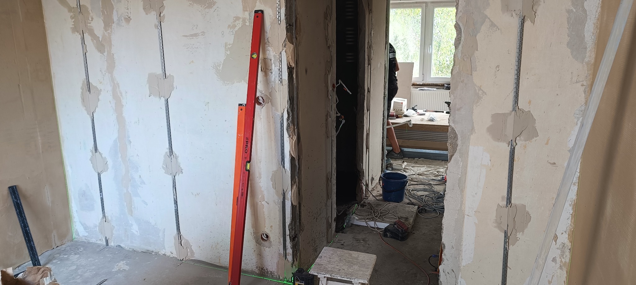 Montaż profili do płyt gipsowo-kartonowych na ścianie z widocznymi łatami szpachli, poziomice, oraz narzędzia w tle. Prace remontowe w trakcie realizacji.