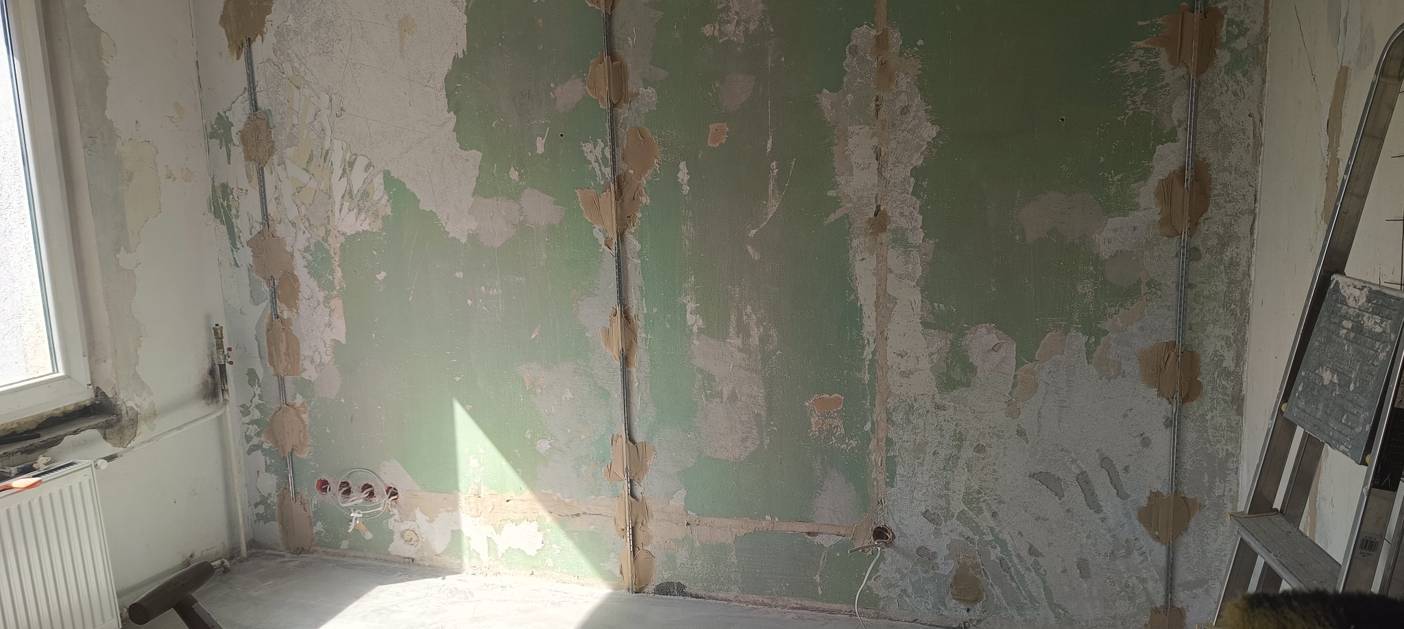 Ściana w trakcie przygotowania do malowania: widoczne ślady po szpachlowaniu, starej farbie i instalacji elektrycznej. W rogu stoi drabina.