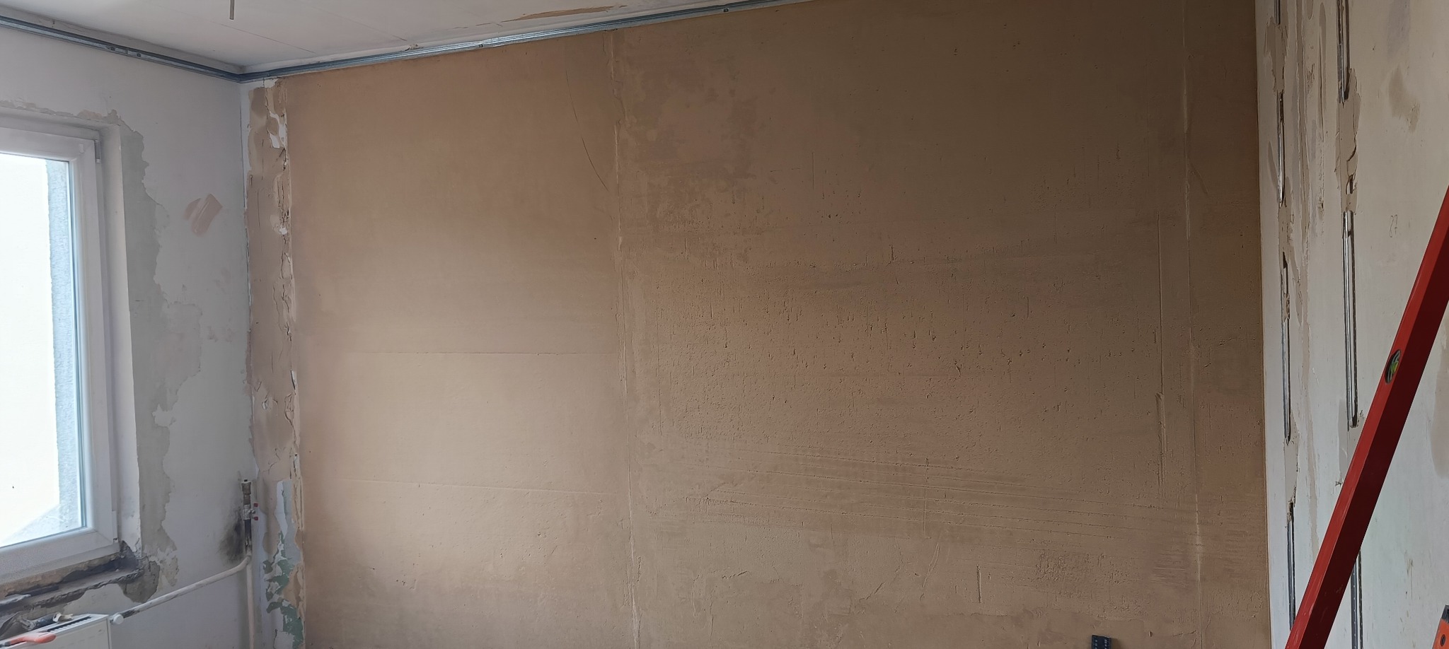 Ściana po nałożeniu szpachli w trakcie remontu, widoczne profile montażowe i poziomica. Fragment okna po lewej stronie kadru.
