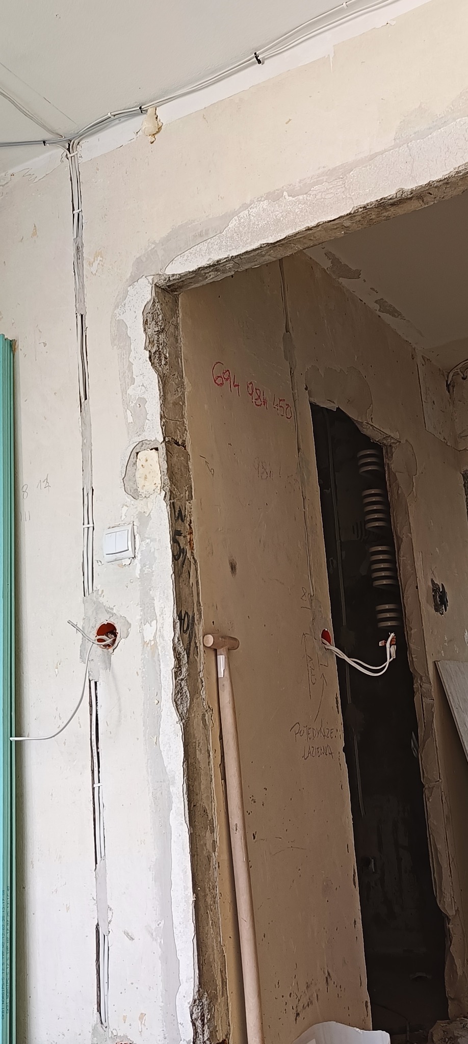 Ściana w trakcie remontu z widocznymi przewodami elektrycznymi, uszkodzonym tynkiem i numerem telefonu napisanym na ścianie przy framudze drzwi.