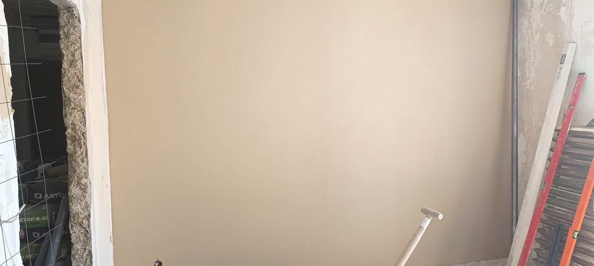 Gładka, beżowa ściana po szpachlowaniu, widoczne narzędzia i materiały budowlane, fragment otworu drzwiowego w trakcie remontu.