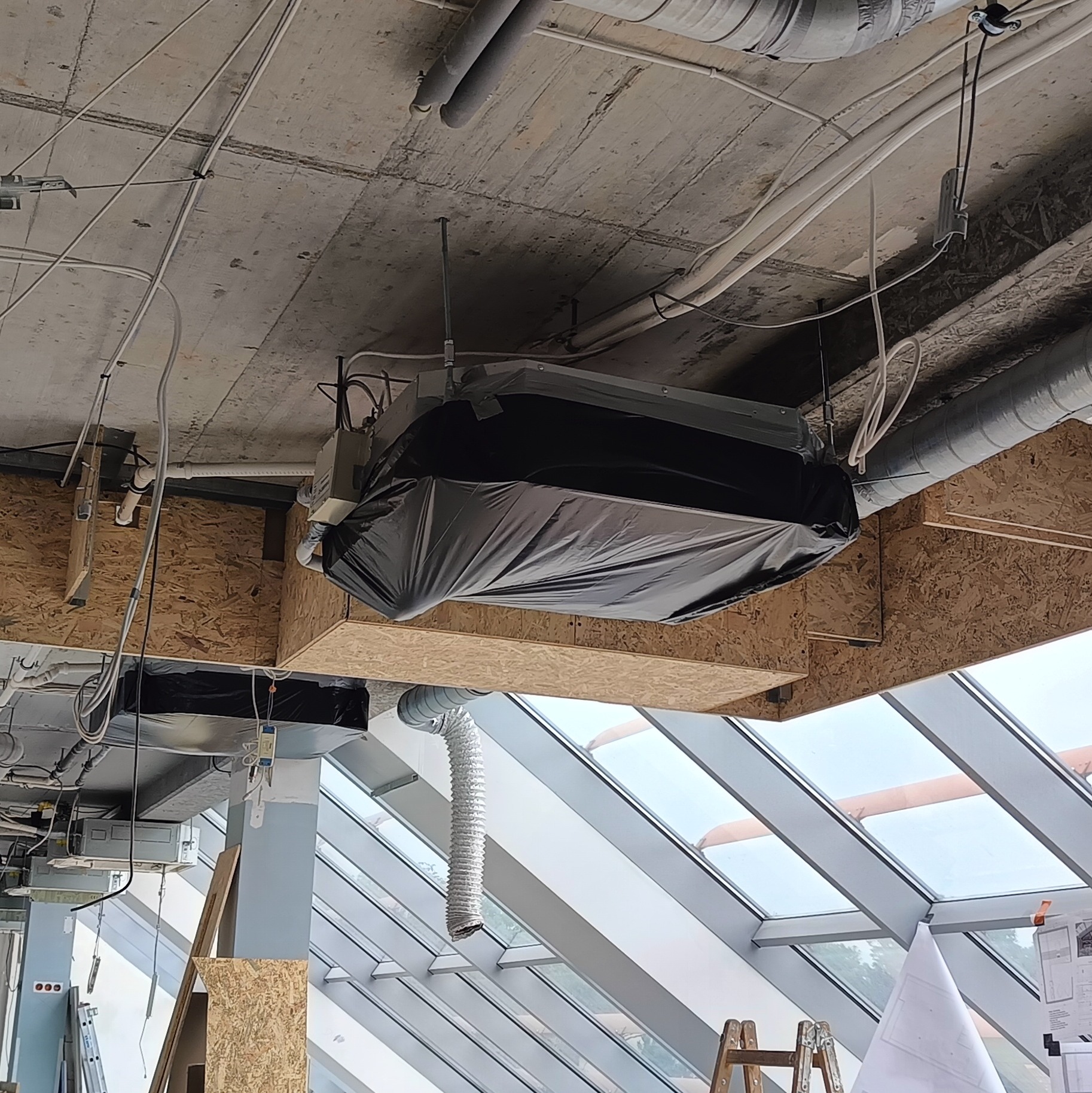 Klimatyzator osłonięty folią podczas prac wykończeniowych w jasnym pomieszczeniu z przeszklonym dachem. Widoczne rury wentylacyjne i kable pod sufitem.