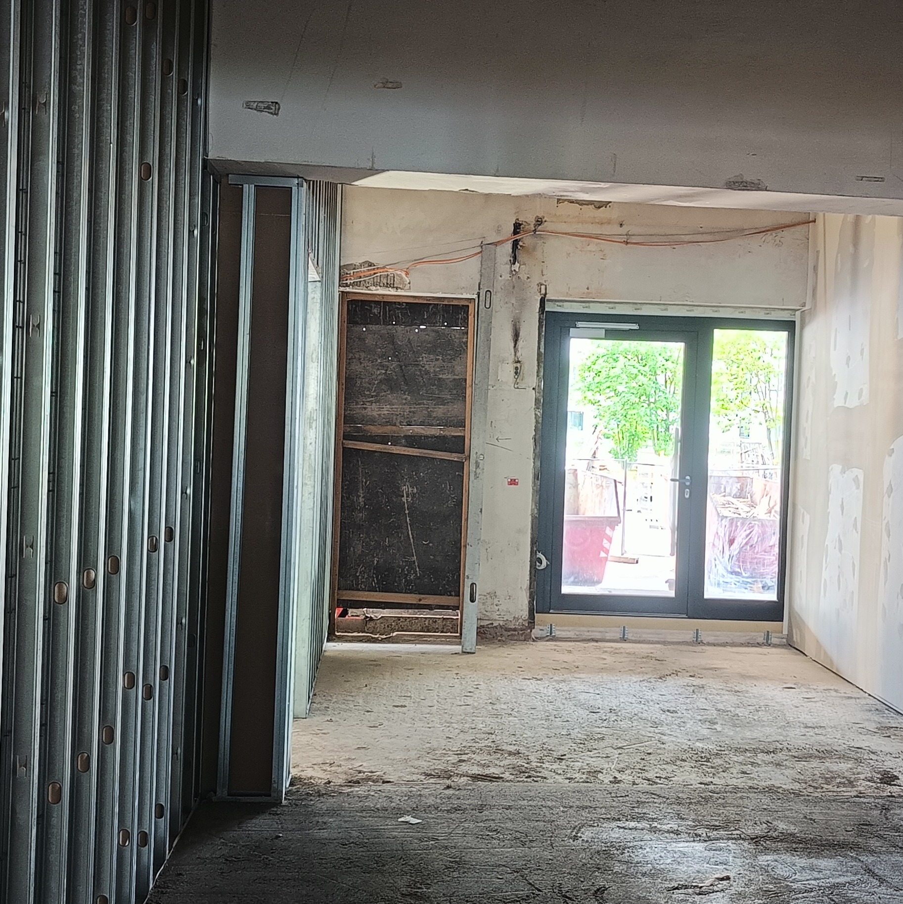 Wnętrze w trakcie prac wykończeniowych: widoczne profile metalowe, ściany przygotowane do malowania, nowe okno balkonowe. Elementy instalacji elektrycznej na ścianie.