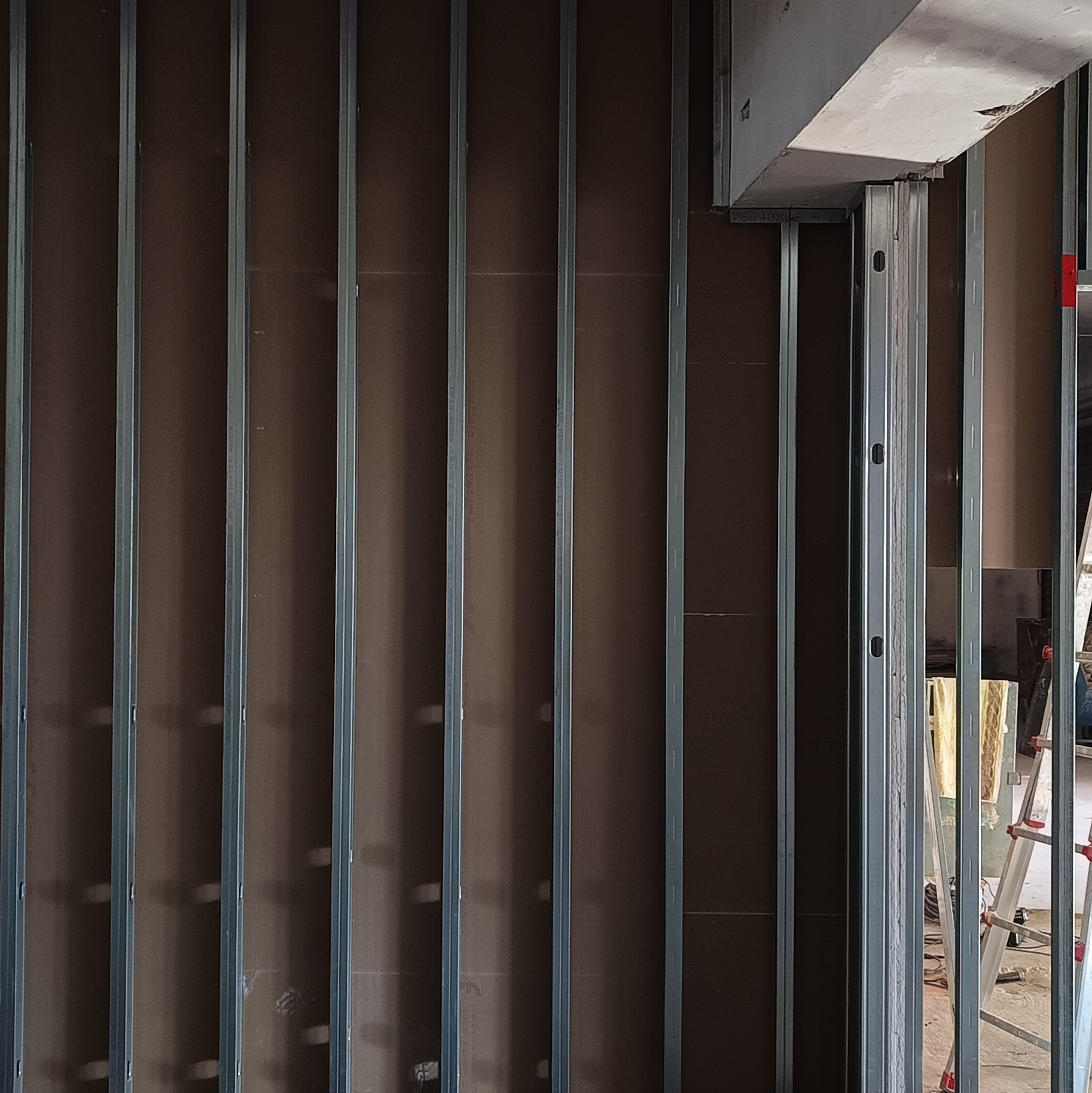 Konstrukcja z profili metalowych i płyty K-G na ścianie, widoczna drabina i materiały izolacyjne w tle. Etap prac wykończeniowych, przygotowanie pod malowanie.