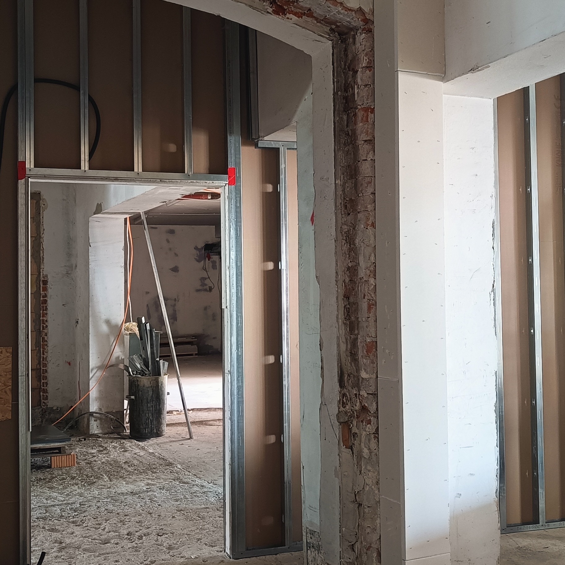 Montaż ścianki działowej z płyty karton-gips: metalowy stelaż z czerwoną taśmą, widoczne instalacje wewnątrz, surowa cegła w tle, etap prac wykończeniowych.