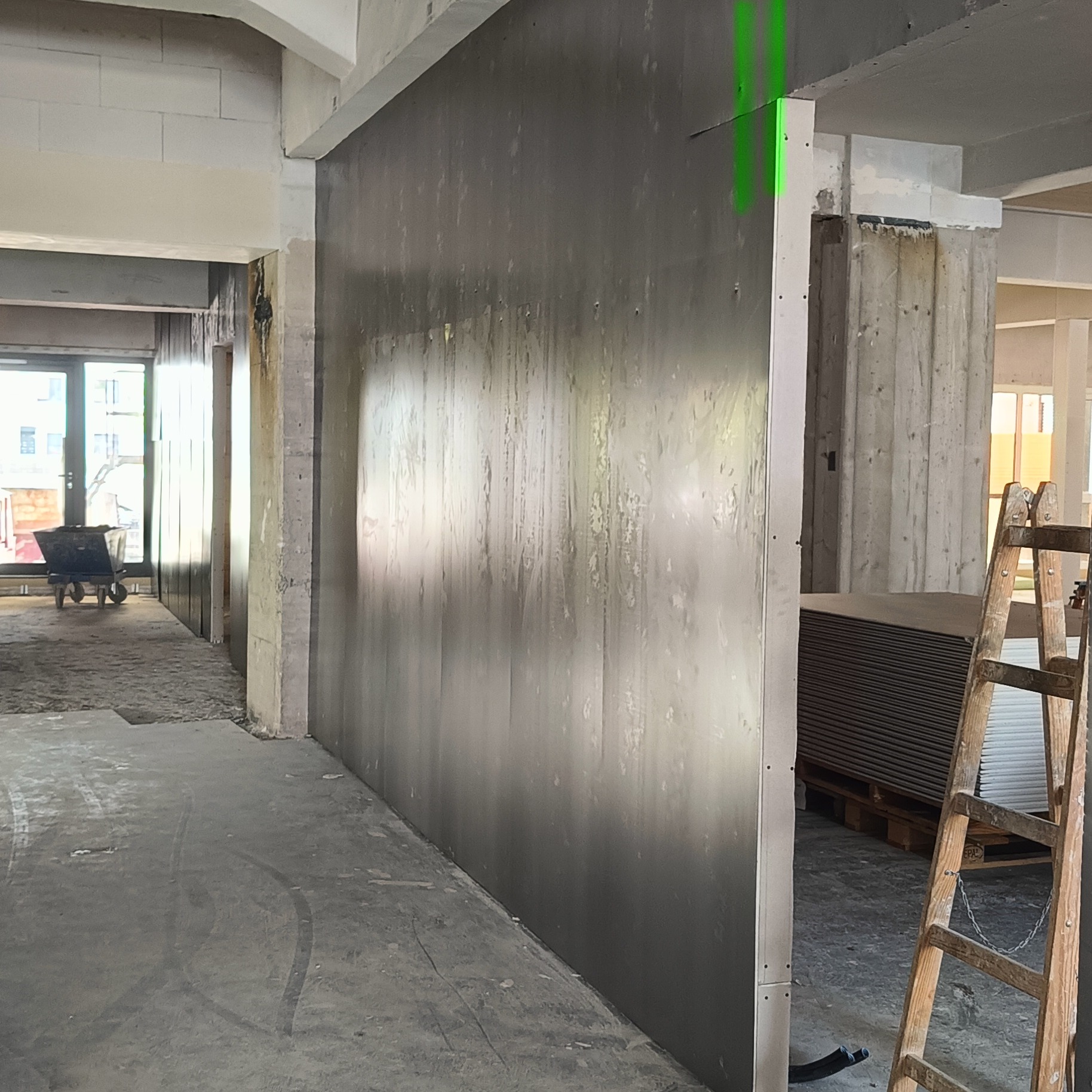 Montaż ścian działowych z płyty karton-gips w surowym wnętrzu. Widoczne płyty na palecie i drabina. Ściana z zielonym oznaczeniem pionu.