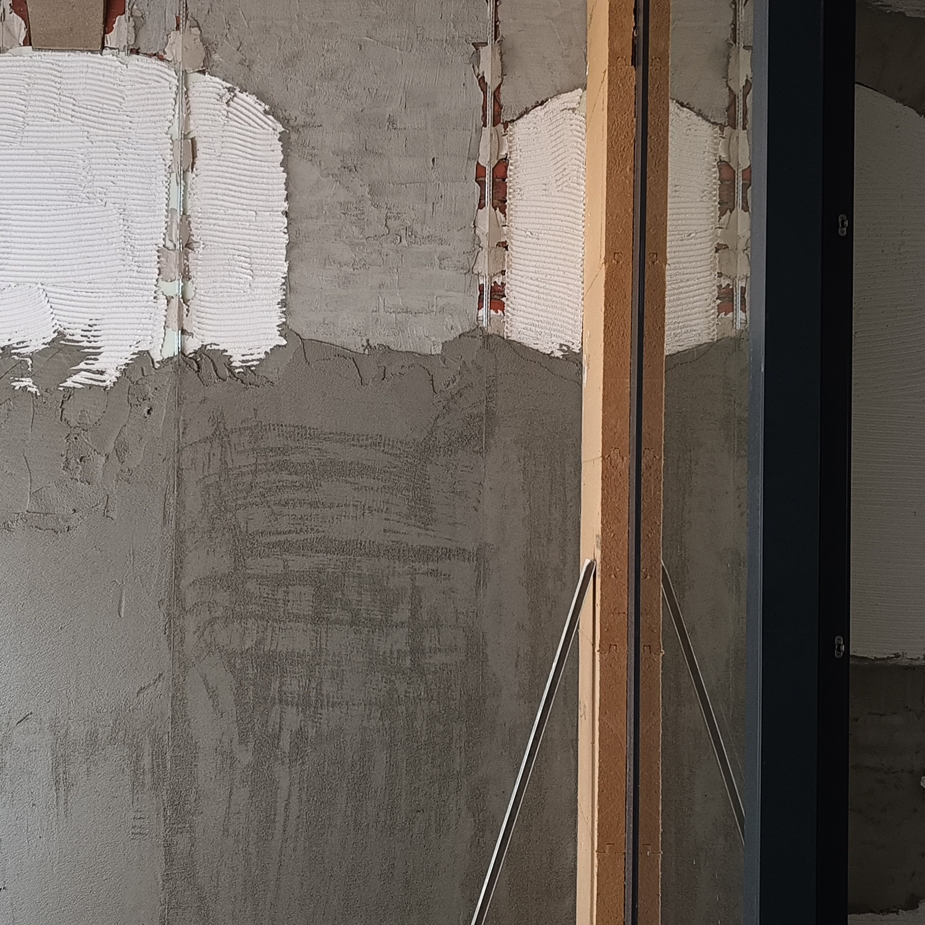 Ściana w trakcie prac wykończeniowych: widoczne fragmenty starej cegły, nałożona warstwa szpachli i przygotowanie podłoża pod dalsze prace, rama okienna z boku.