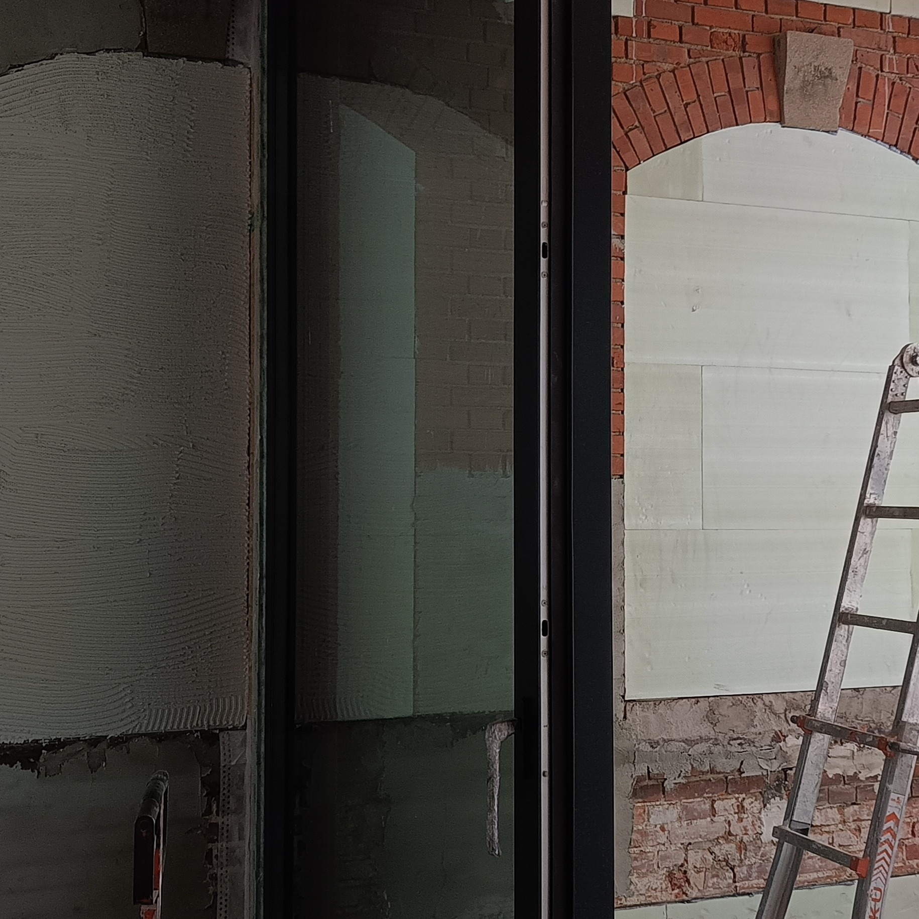 Prace wykończeniowe w Krakowie: ściana z nałożoną warstwą szpachli, okno z czarną ramą i fragment drabiny. Widoczny fragment ceglanego łuku.