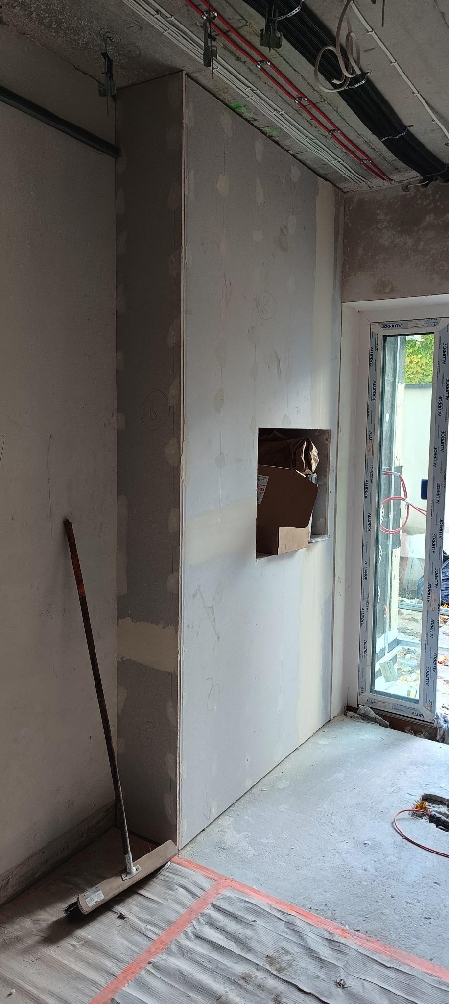 Ściana z gładzią gipsową w trakcie prac wykończeniowych. Widoczne instalacje pod sufitem, okno balkonowe i narzędzia. W ścianie otwór na dekoracyjny element.