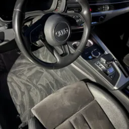 Wnętrze Audi A4: czarna kierownica, deska rozdzielcza, automatyczna skrzynia bieg&oacute;w i wyprana, ciemnoszara tapicerka fotela kierowcy z wyraźnym wzorem.