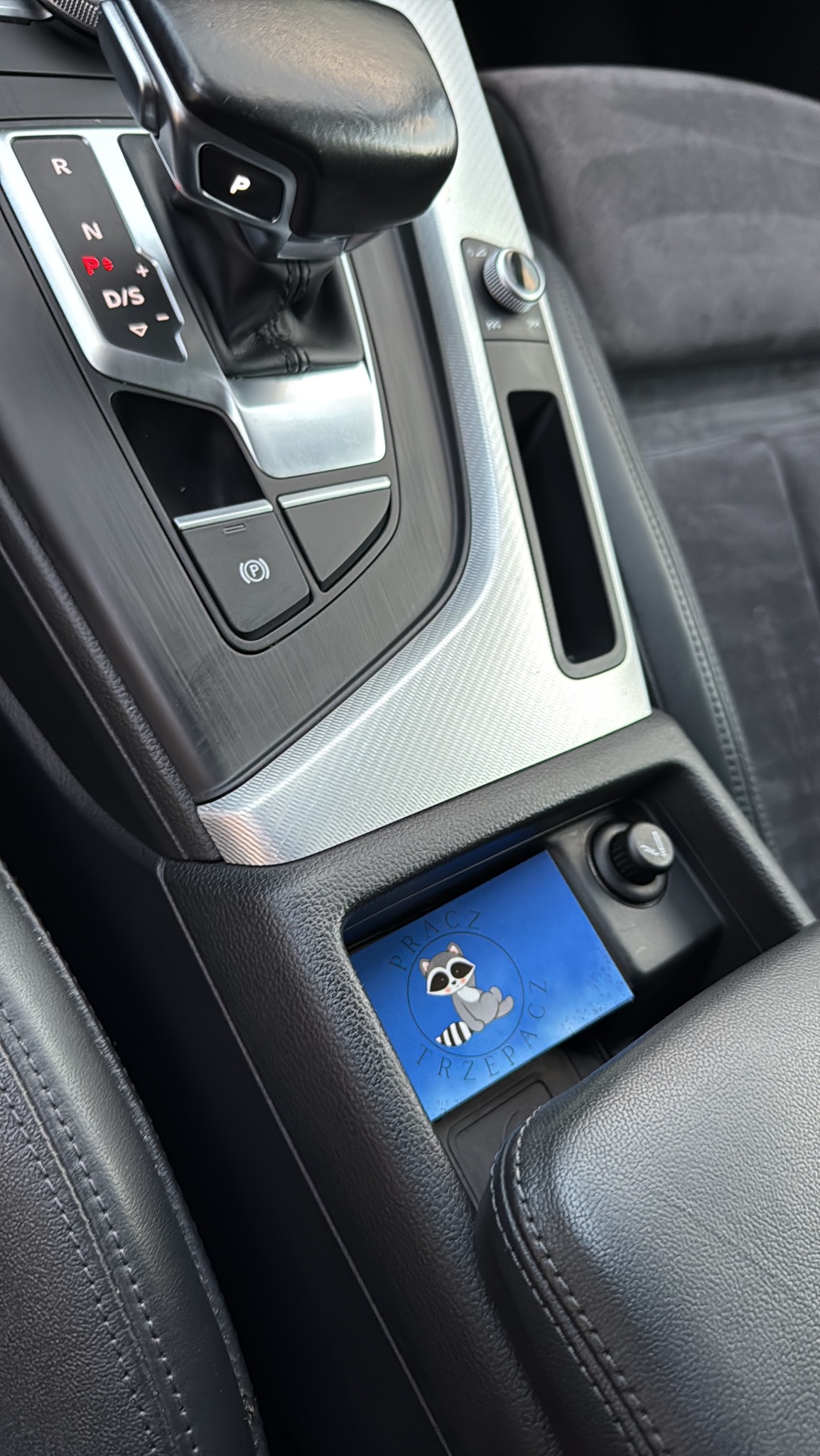 Wnętrze auta z logo firmy czyszczącej tapicerkę. Widoczny fragment deski rozdzielczej, skórzane fotele i logo z szopem praczem. Detale czyszczenia tapicerki samochodowej.