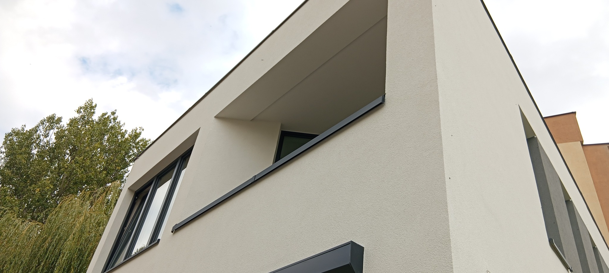 Nowoczesny budynek z balkonem i oknami o czarnych ramach, widziany z dołu pod kątem. Jasna elewacja, minimalistyczny design, fragment drzewa w tle.