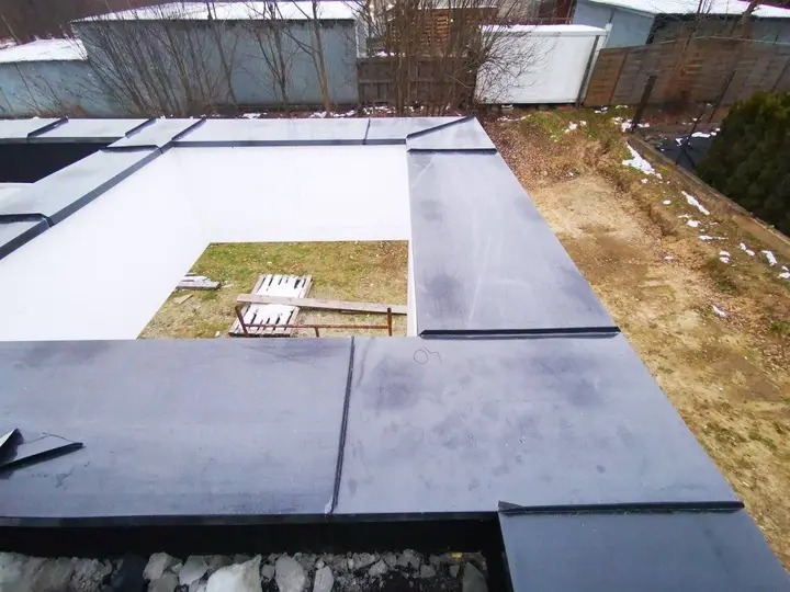 Attyka wykończona na felc podwójny, widok z góry. Szare obróbki blacharskie na tle białej ściany i zielonego trawnika. Fragment dachu z widocznymi łączeniami.