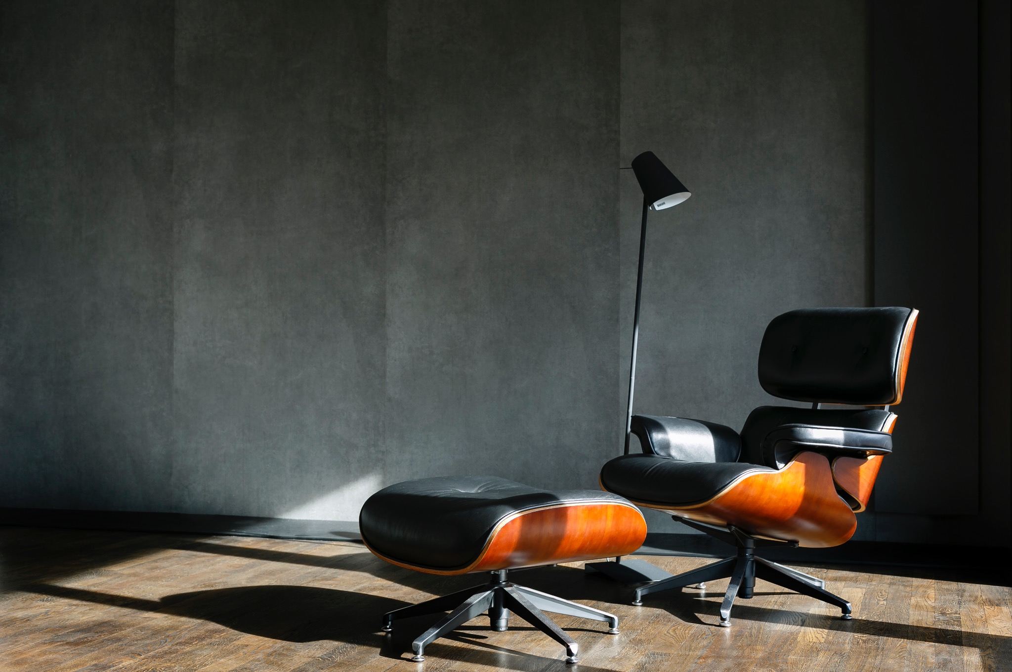 Eames Lounge Chair z podnóżkiem i lampą na tle szarej ściany. Parkiet odbija światło słoneczne. Minimalistyczny design i elegancja.