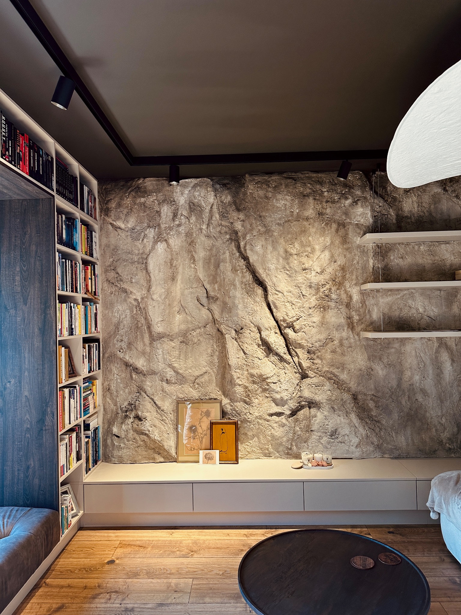 Dekoracyjna ściana imitująca skałę w salonie z oświetleniem punktowym, półkami i biblioteczką. Wykończenie wnętrza w nowoczesnym stylu z elementami naturalnymi.