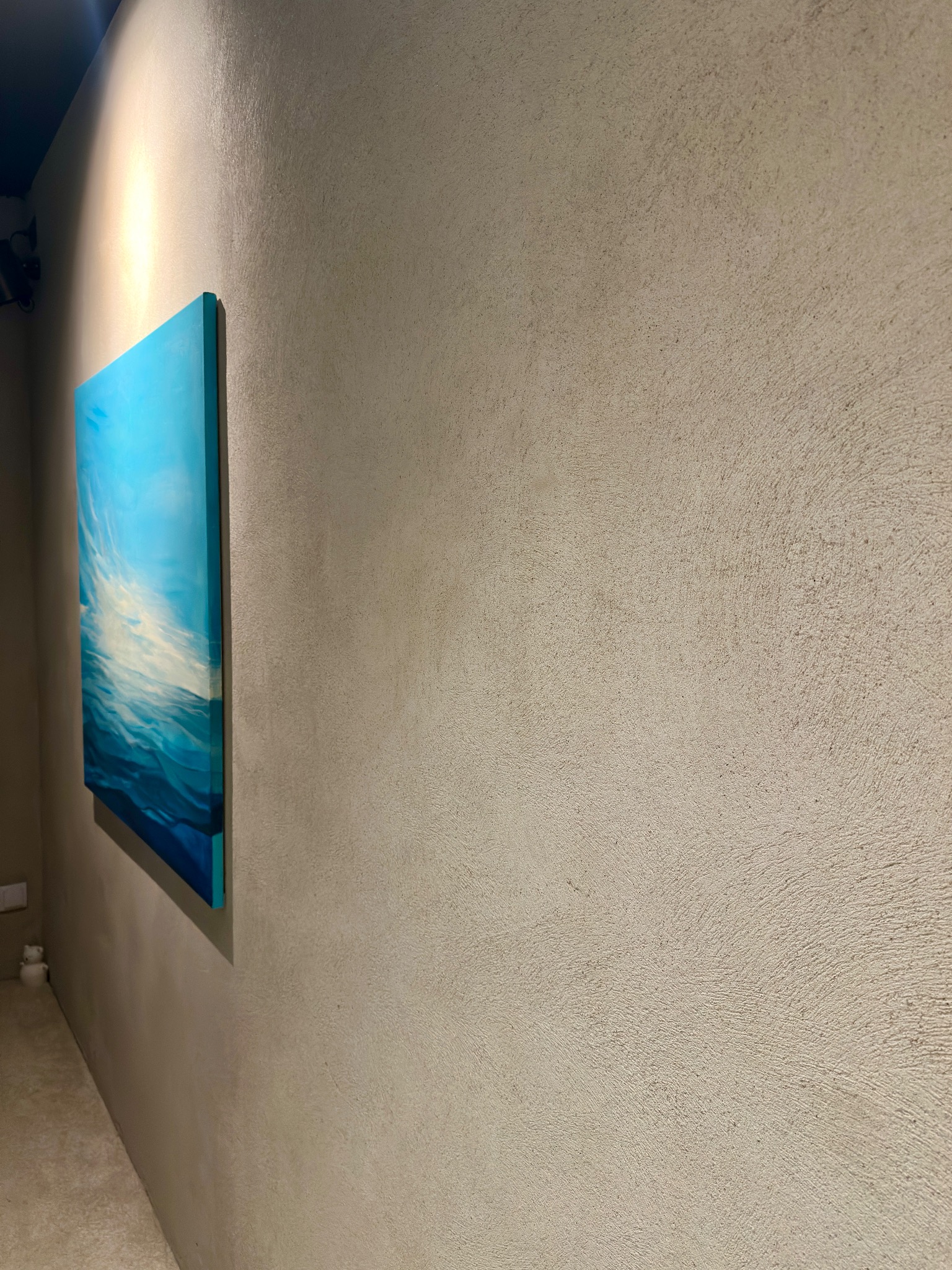 Ściana z dekoracyjną, beżową fakturą i zawieszonym obrazem z motywem morza, oświetlona punktowym światłem. Widoczny fragment podłogi i gniazdko.