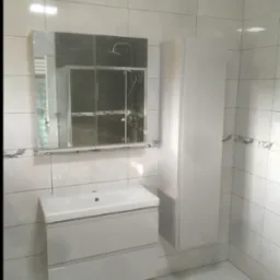 Biała umywalka z szafką, lustro i wysoka szafka w wykafelkowanej łazience. Widoczne płytki podłogowe i ścienne w odcieniach bieli i szarości.