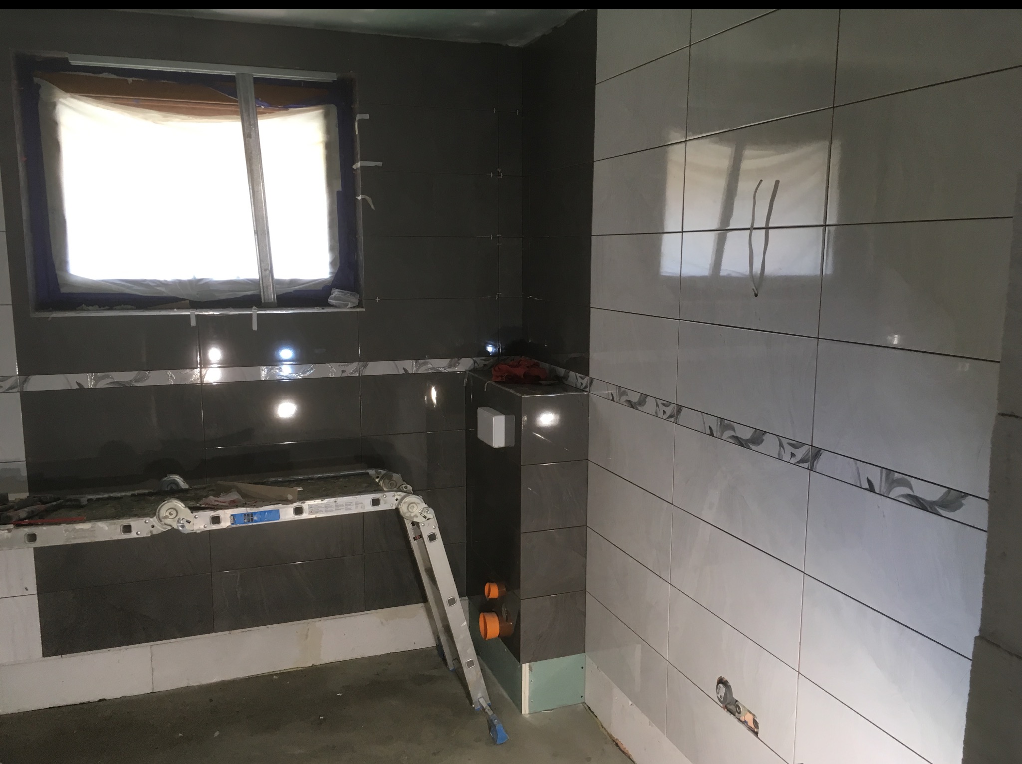 Wnętrze łazienki w trakcie prac wykończeniowych: częściowo ułożone płytki w odcieniach szarości i bieli, drabina, okno zabezpieczone folią. Widoczne elementy instalacji.