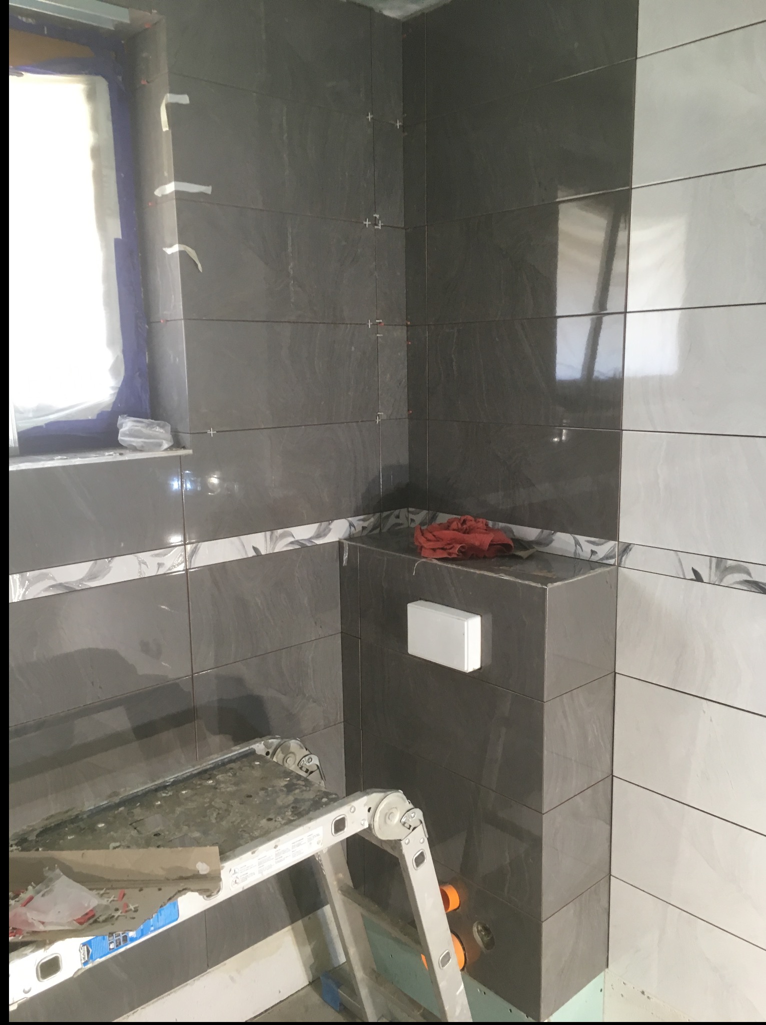 Wnętrze łazienki w trakcie prac glazurniczych. Widoczne szare i białe płytki na ścianach, drabina malarska i wystające rury. W tle okno z zasłoniętym widokiem.