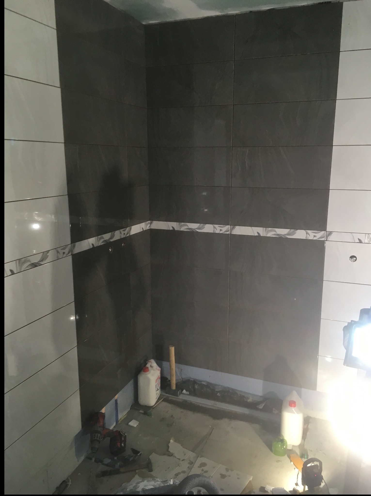 Łazienka w trakcie remontu: ściany wyłożone szarymi i białymi płytkami z dekoracyjnym pasem. Na podłodze widoczne narzędzia i materiały budowlane.