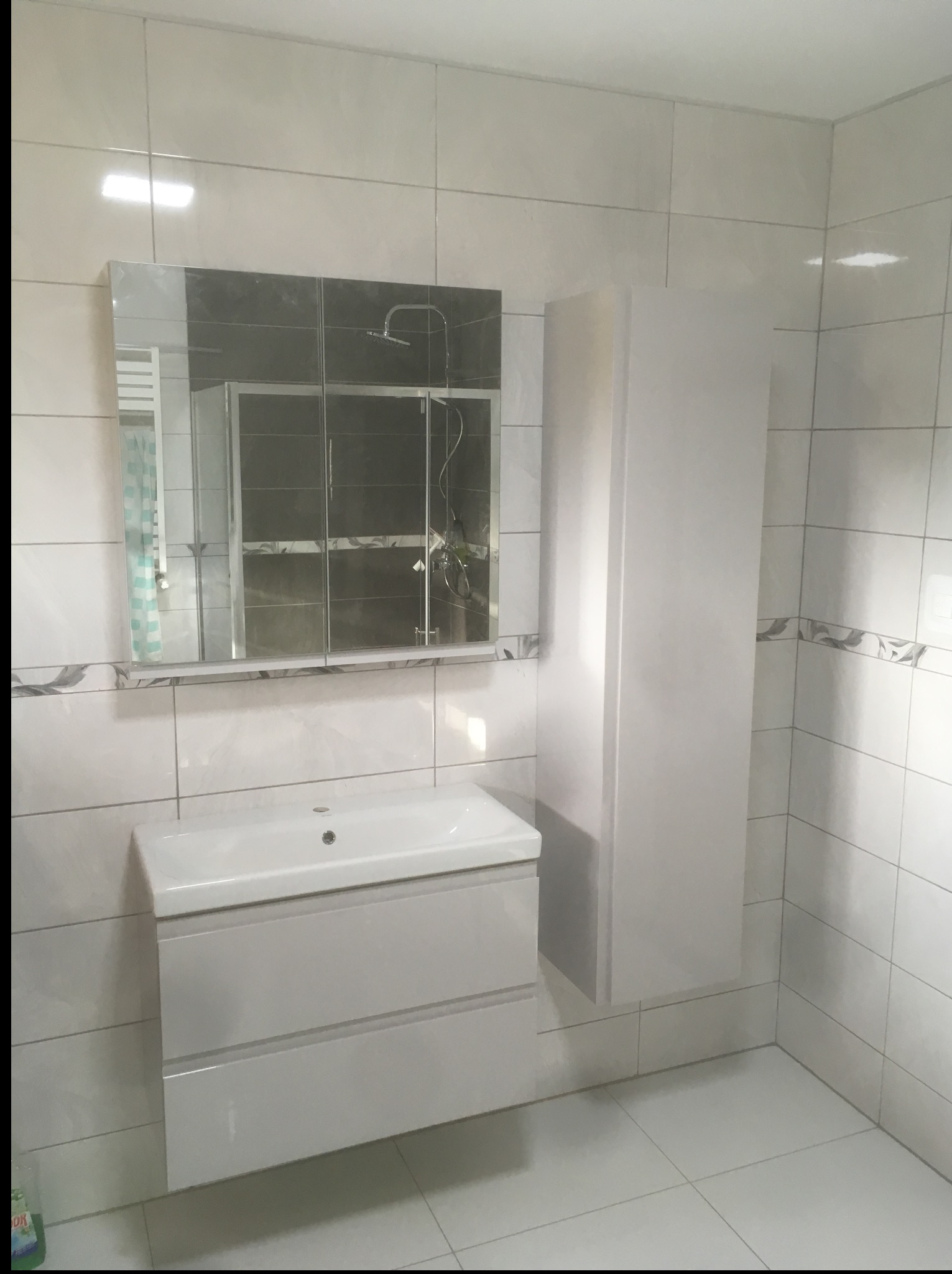 Biała umywalka z szafką, lustro i wysoka szafka w wykafelkowanej łazience. Widoczne płytki podłogowe i ścienne w odcieniach bieli i szarości.