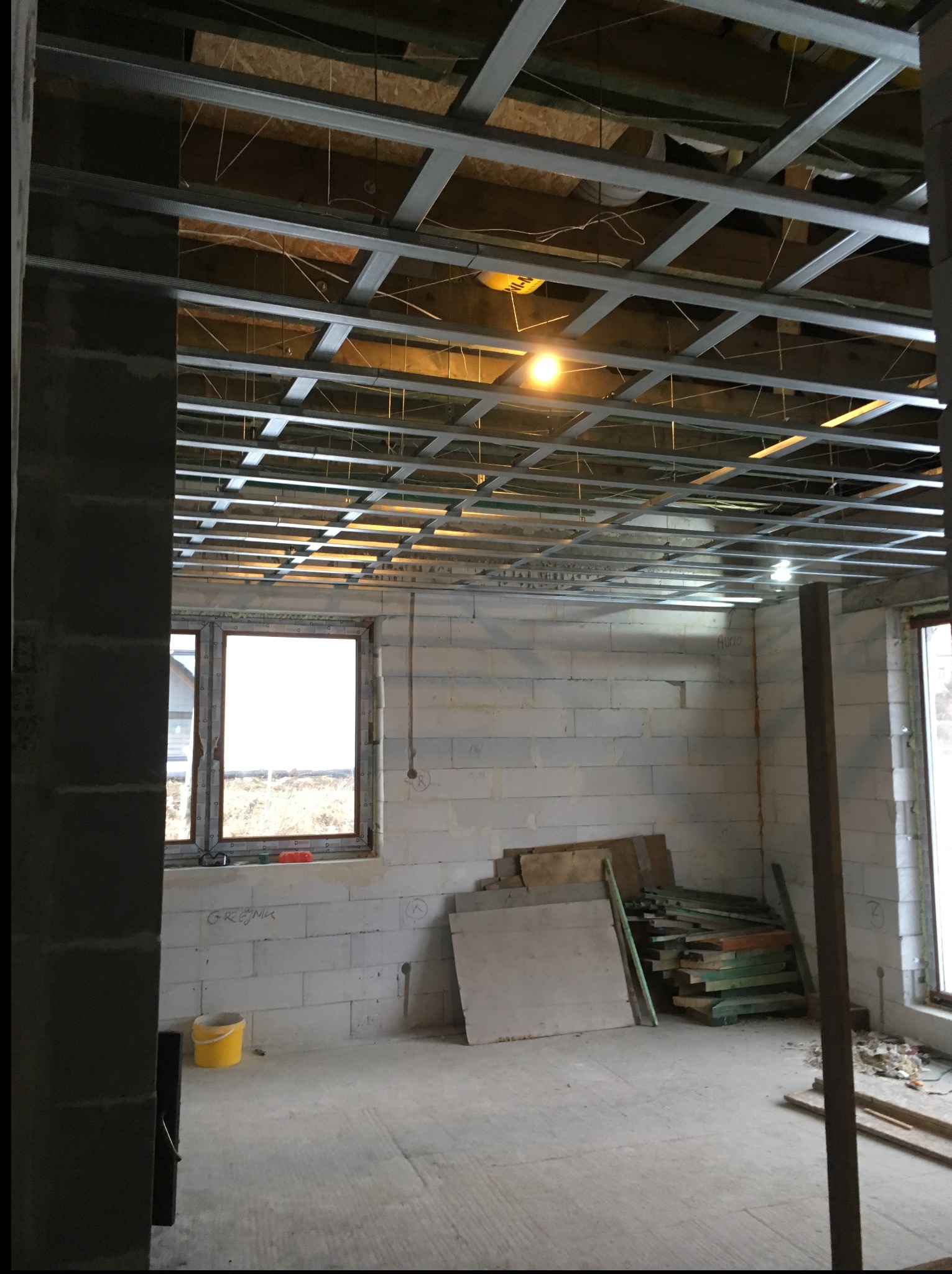 Wnętrze w trakcie prac wykończeniowych: surowa ściana z pustaków, metalowa konstrukcja sufitu podwieszanego, okno z widokiem na zewnątrz, narzędzia pracy.