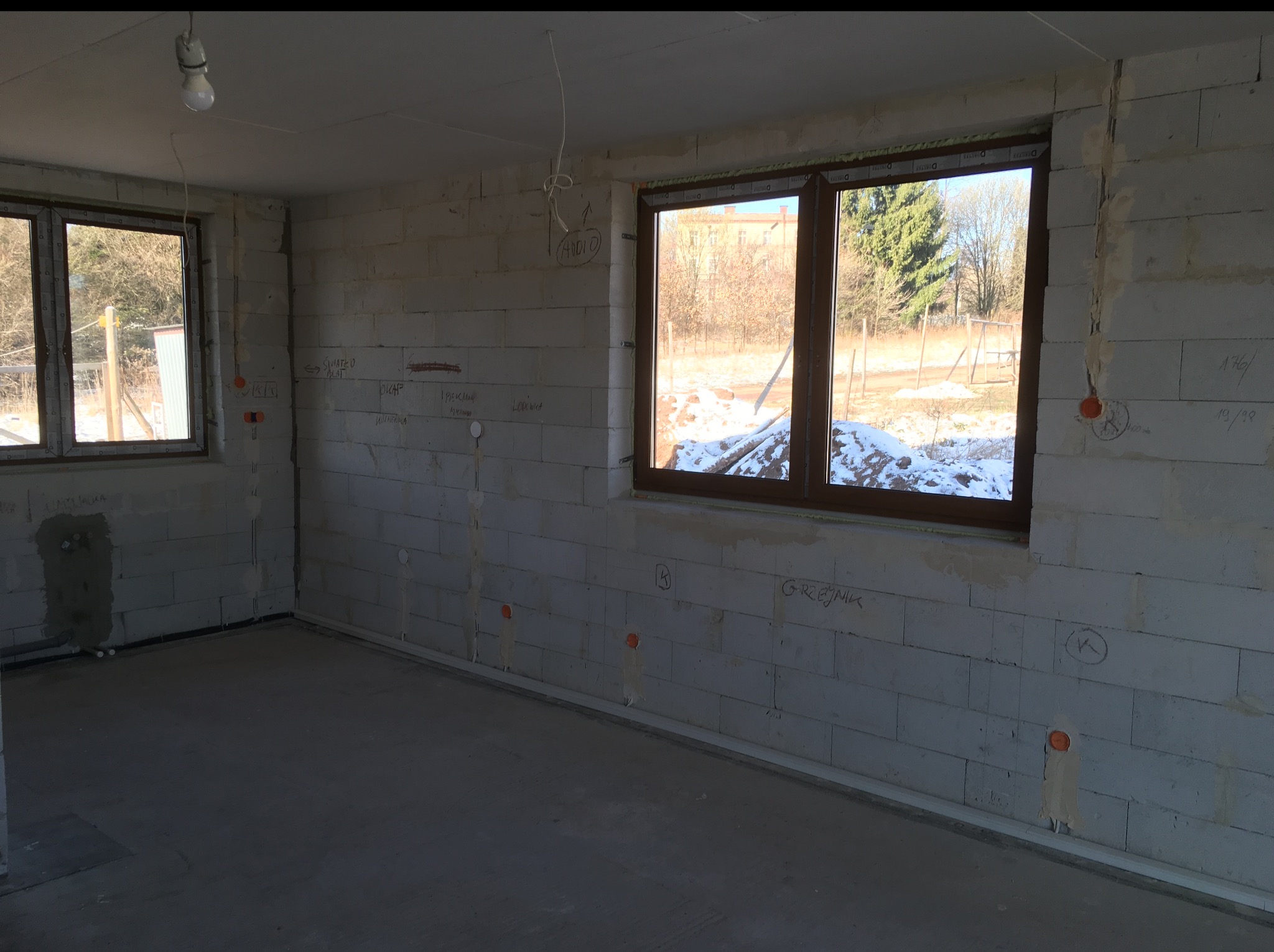 Surowy stan wnętrza z bloków, widoczne okna, instalacje elektryczne i oznaczenia na ścianach. Podłoga betonowa, sufit z wiszącą żarówką. Widok z okna na otoczenie.