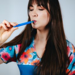 LOGOSPOT Terapia logopedyczna i miofunkcjonalna Martyna Majewska - Kobieta w kwiecistej bluzce ćwiczy wymowę za pomocą niebieskiego ustnika. Narzędzie logopedyczne w zbliżeniu. Ćwiczenia artykulacyjne.