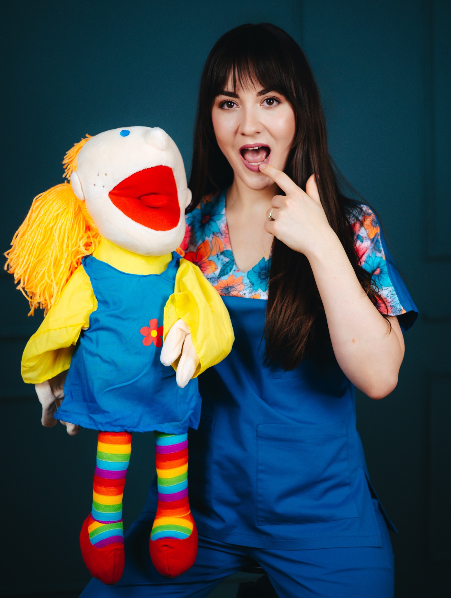 Logopeda prezentuje ćwiczenie z lalką o dużych ustach i tęczowych nogach, demonstrując prawidłową artykulację. Kobieta w niebieskim uniformie pokazuje ułożenie języka.