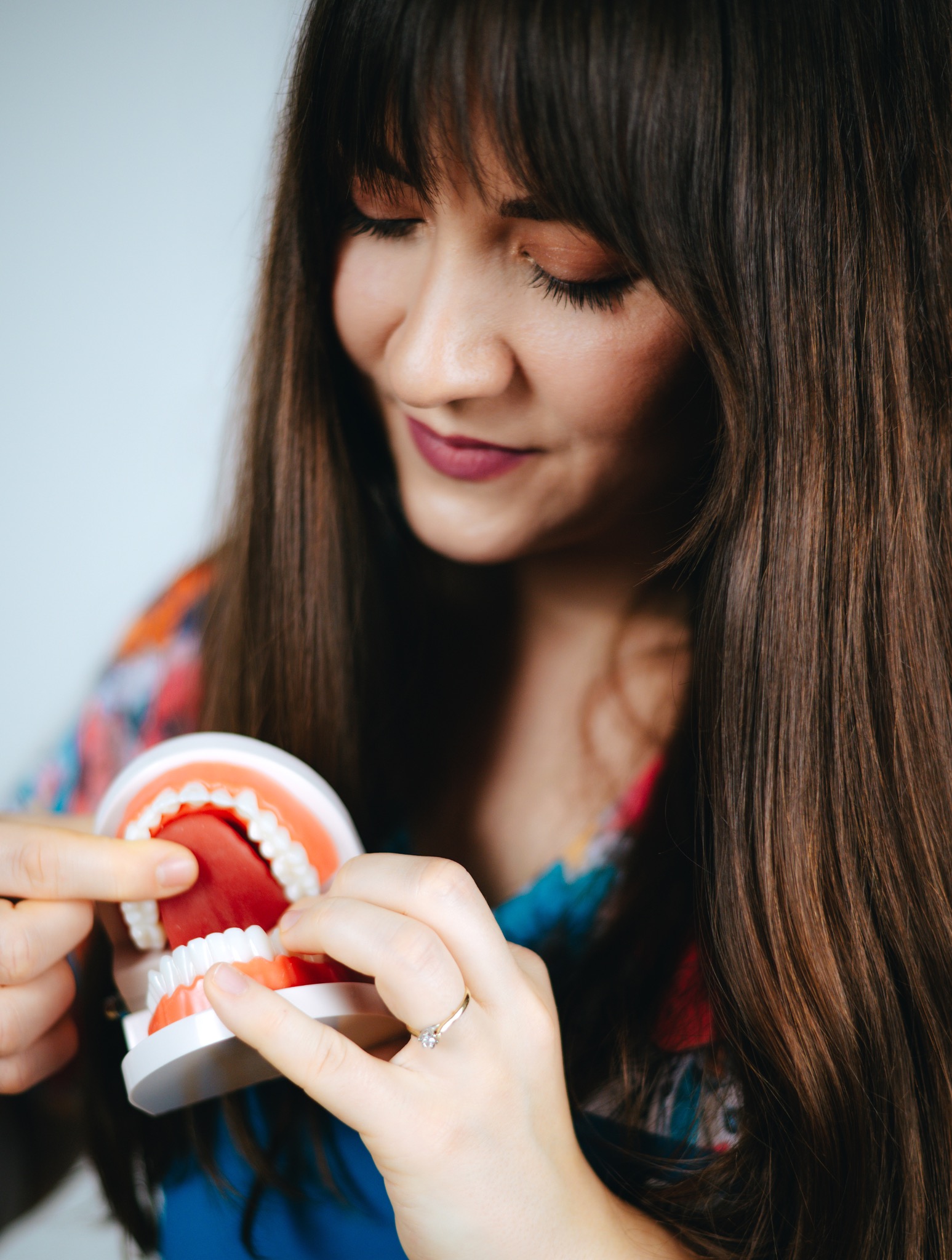 Kobieta z długimi włosami demonstruje model szczęki, wskazując palcem na zęby. Model w jasnych kolorach, dłoń z pierścionkiem. Nauka poprawnej wymowy.