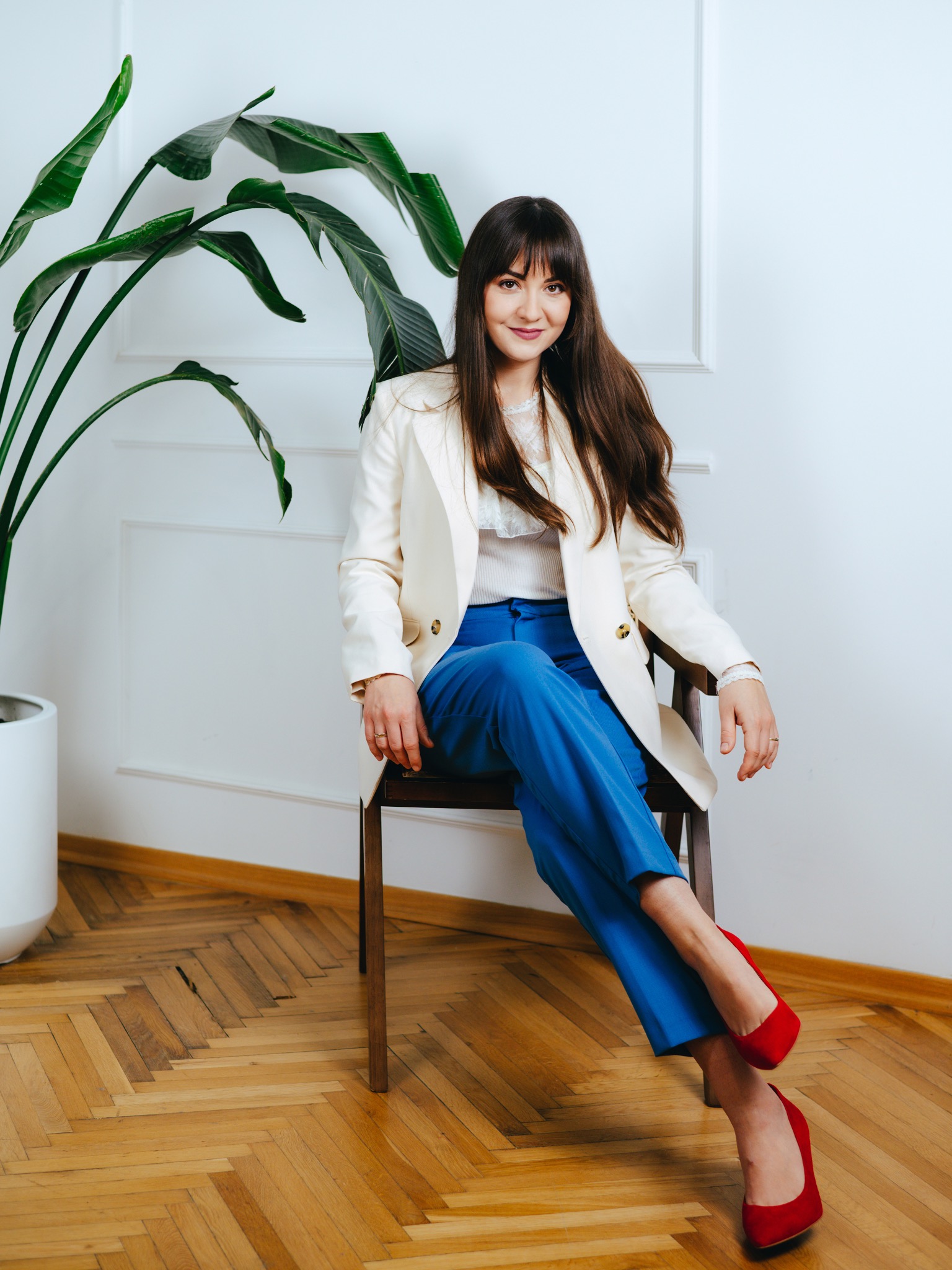 Kobieta siedzi na krześle, ubrana w białą marynarkę, niebieskie spodnie i czerwone buty, z zieloną rośliną w tle. Uśmiech na twarzy, ciemne włosy.