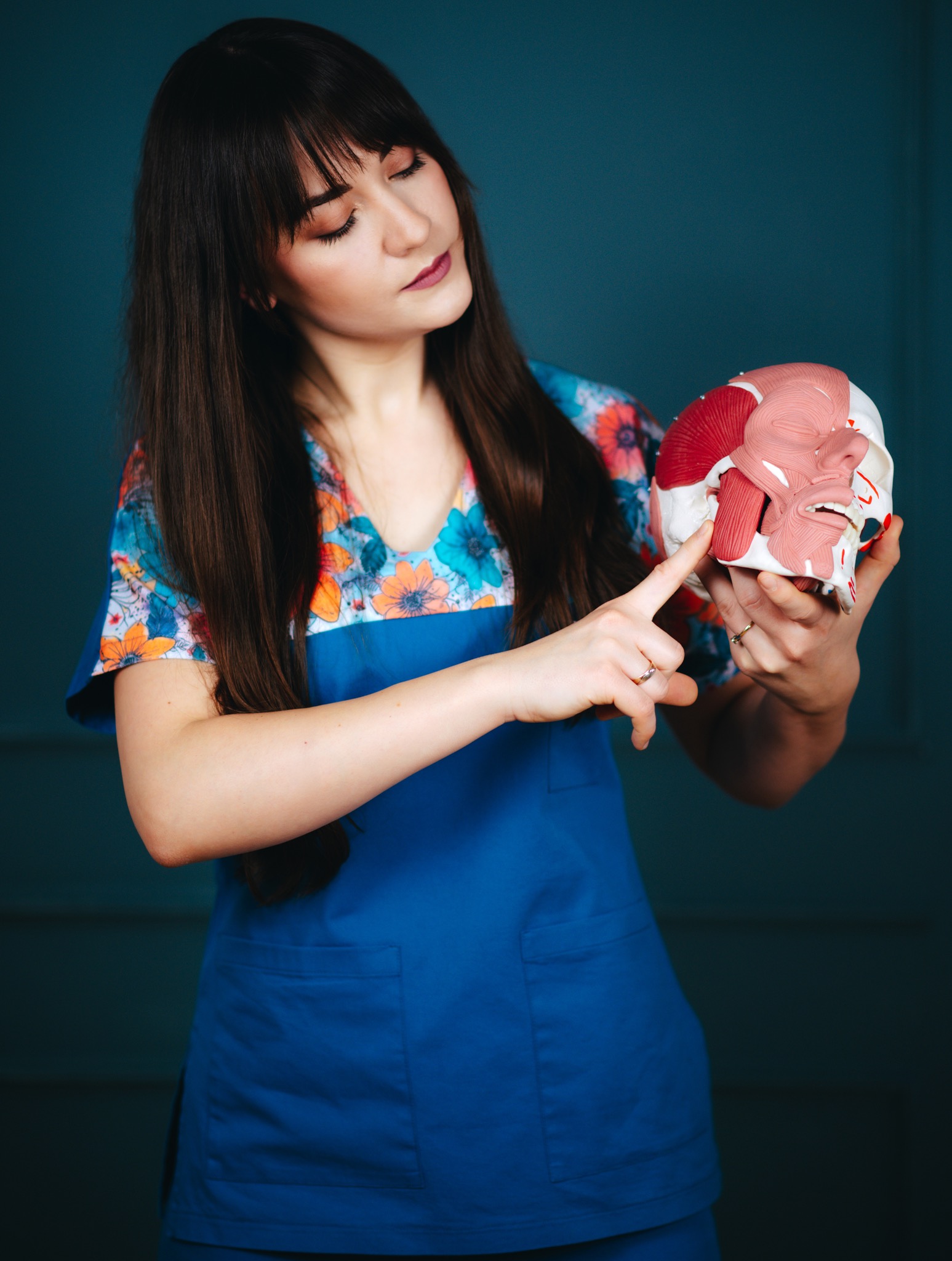 Kobieta w niebieskim uniformie wskazuje na model czaszki z mięśniami twarzy. Prezentacja anatomii w gabinecie lekarskim na tle ciemnej ściany.