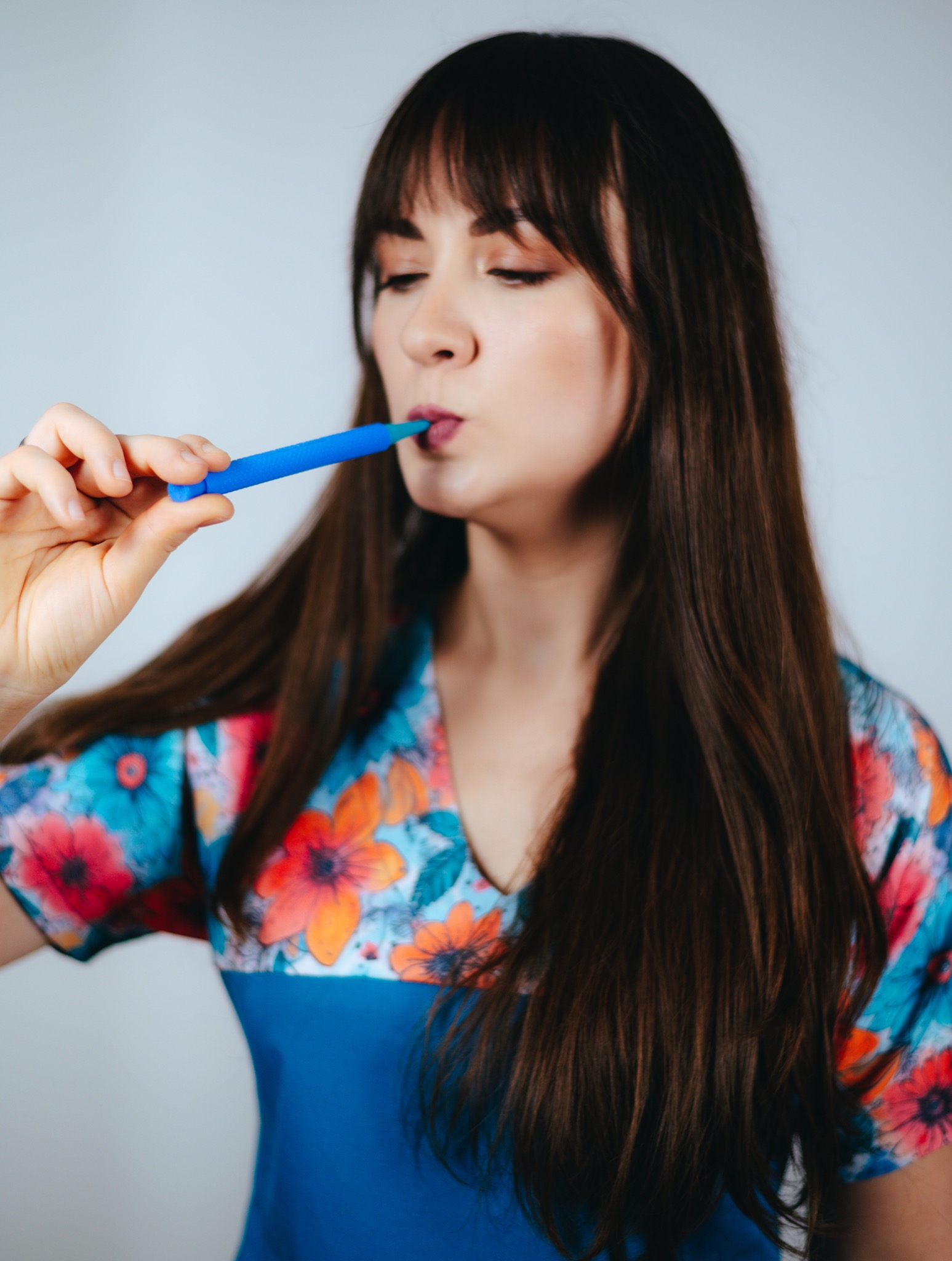 Kobieta w kwiecistej bluzce ćwiczy wymowę za pomocą niebieskiego ustnika. Narzędzie logopedyczne w zbliżeniu. Ćwiczenia artykulacyjne.