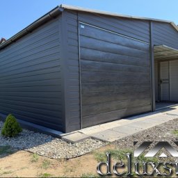 DELUXSTAL Grzegorz Rus - Grafitowy garaż z blachy drewnopodobnej, z otwartymi drzwiami i zadaszeniem, otoczony kamieniami i zielenią. Widoczny system rynnowy i fragment domu w tle.