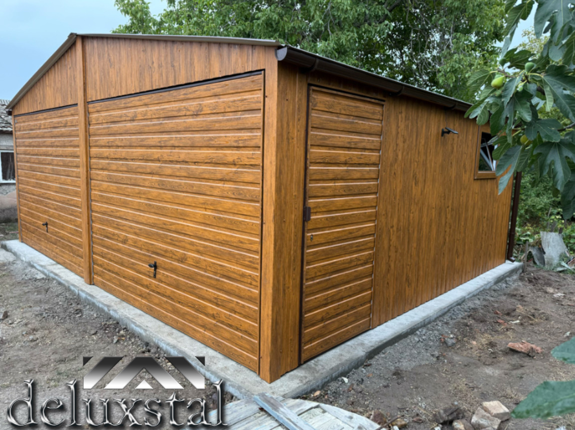 Brązowy garaż blaszany z dwiema bramami segmentowymi i drzwiami wejściowymi z boku, na betonowej wylewce. W tle drzewa i krzewy.