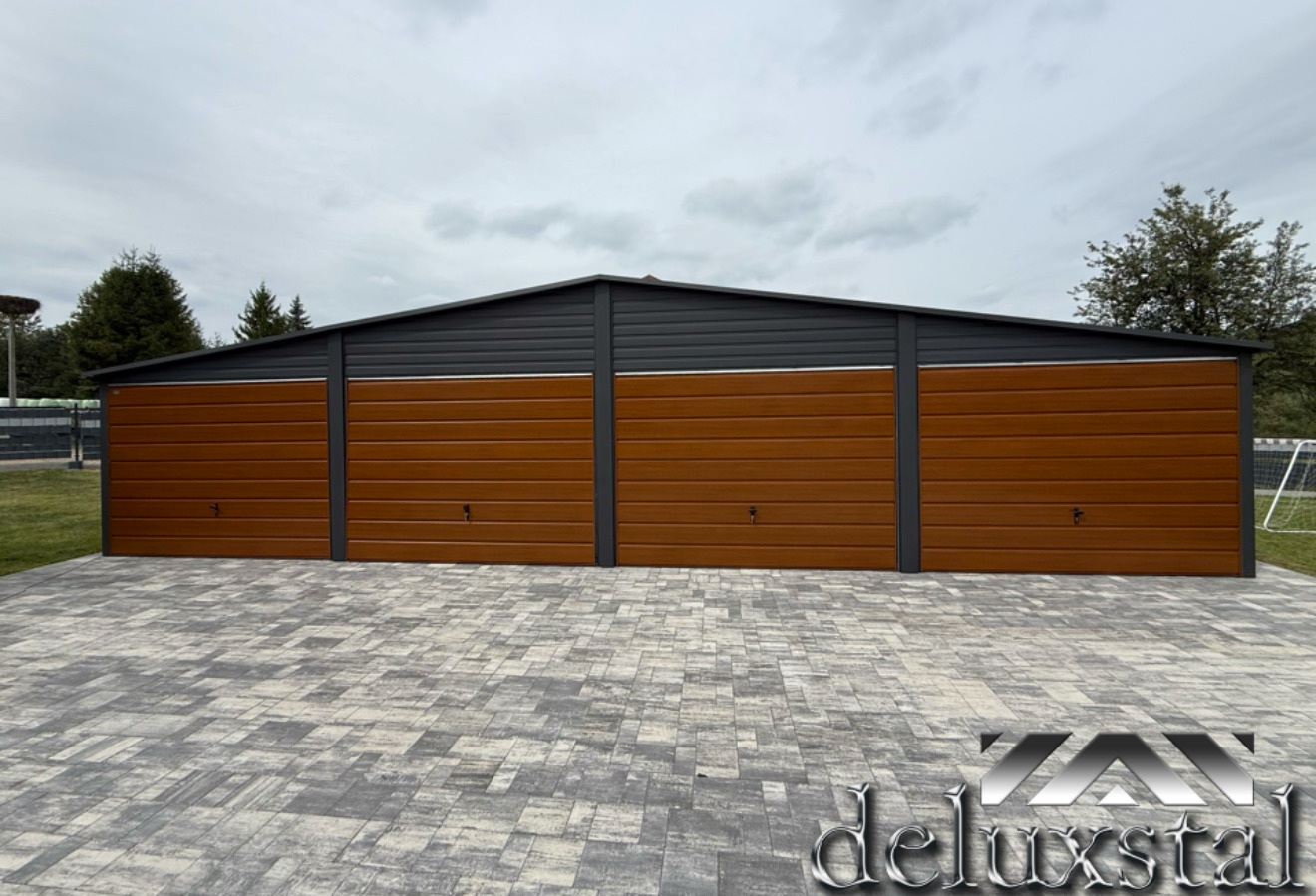 Czterostanowiskowy garaż blaszany z brązowymi bramami segmentowymi i szarą konstrukcją, widoczny z zewnątrz na tle pochmurnego nieba.