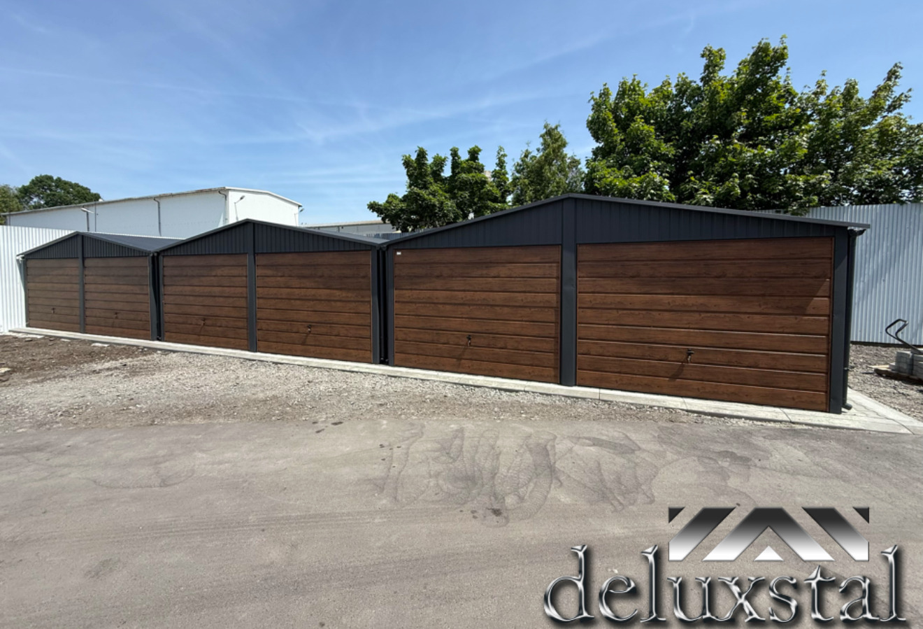 Trzy garaże blaszane z brązowymi bramami segmentowymi i ciemnoszarymi dachami, ustawione w rzędzie na żwirowym podłożu, z logo firmy w prawym dolnym rogu.