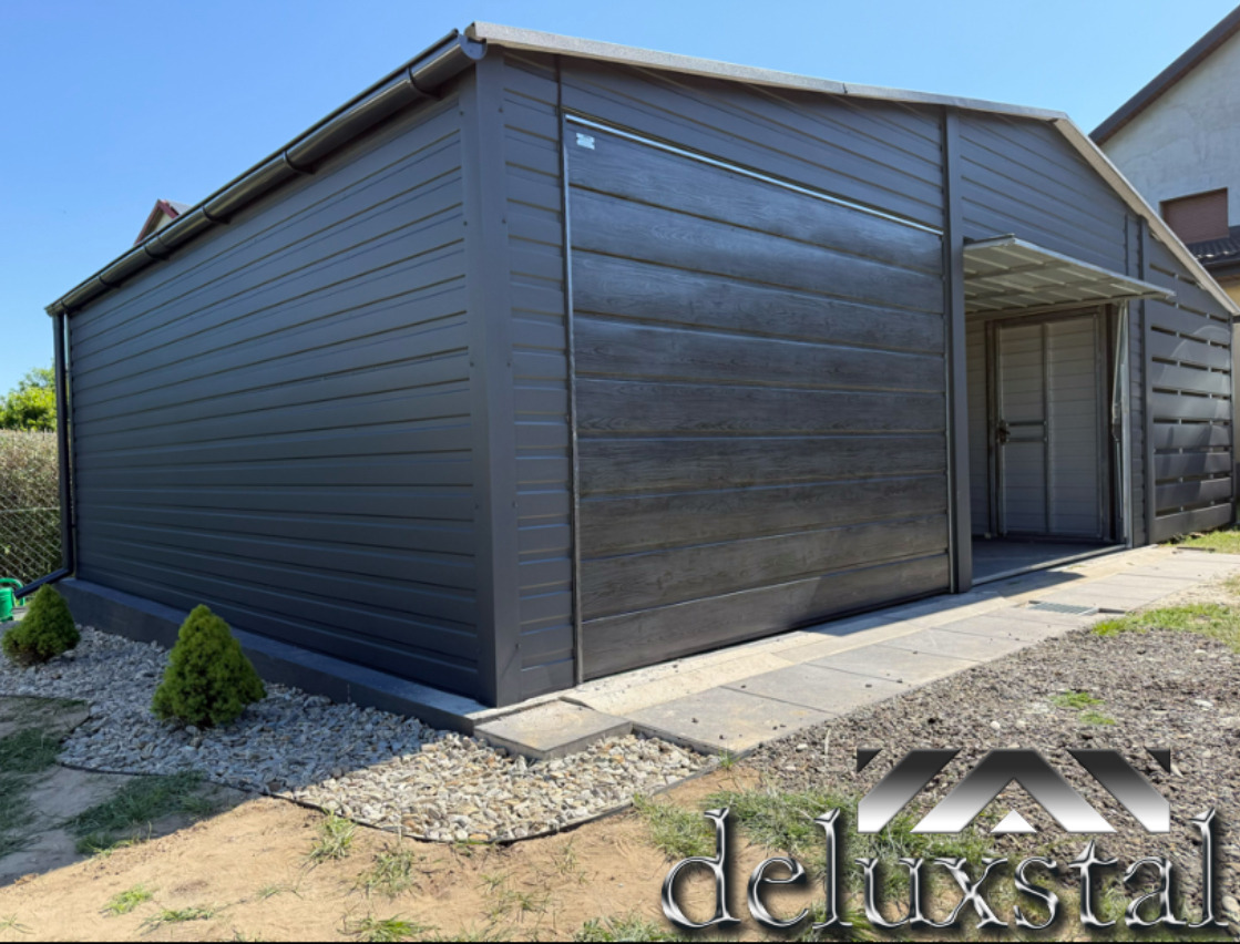 Grafitowy garaż z blachy drewnopodobnej, z otwartymi drzwiami i zadaszeniem, otoczony kamieniami i zielenią. Widoczny system rynnowy i fragment domu w tle.