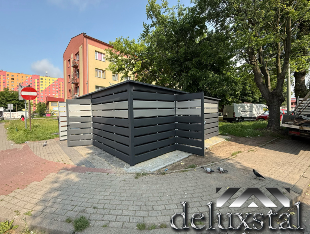 Szary, nowoczesny schowek na śmietniki z poziomymi panelami, otwarty z jednej strony, na tle bloku mieszkalnego i drzew. Widoczne gołębie i logo firmy.