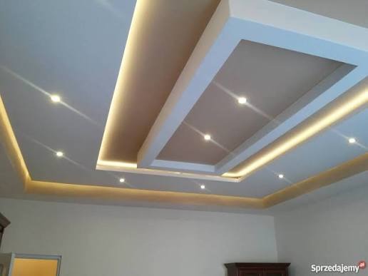 Podwieszany sufit z oświetleniem LED w kształcie prostokątów, widok z dołu. Jasne ściany i fragmenty mebli w tle, ciepłe światło.