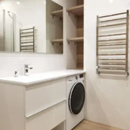 Nowoczesna łazienka z białą szafką umywalkową, pralką w zabudowie, grzejnikiem drabinkowym i drewnianymi półkami. Minimalistyczny design, jasne płytki i podłoga imitująca drewno.