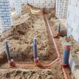 Dobra Atmosfera - Instalacja kanalizacyjna w wykopie ziemnym, pomarańczowe rury z szarymi izolacjami pionowymi, surowe ściany z białego bloczka w tle, widok z góry.