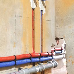 Dobra Atmosfera - Instalacja wodna: rury czerwone i niebieskie prowadzone w ścianie, zabezpieczone uchwytami. Szare rury kanalizacyjne na dole. Surowy tynk w tle.