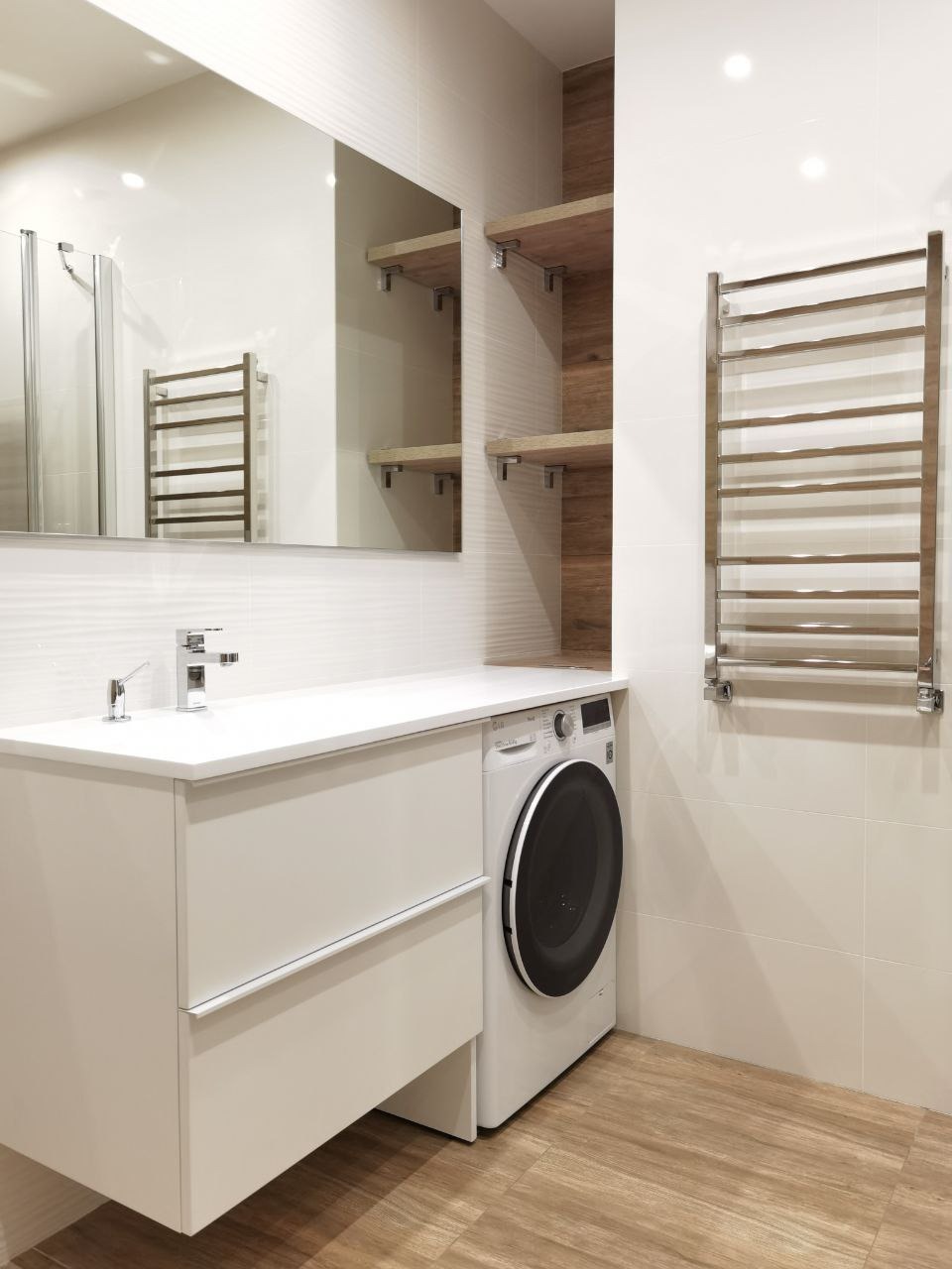 Nowoczesna łazienka z białą szafką umywalkową, pralką w zabudowie, grzejnikiem drabinkowym i drewnianymi półkami. Minimalistyczny design, jasne płytki i podłoga imitująca drewno.