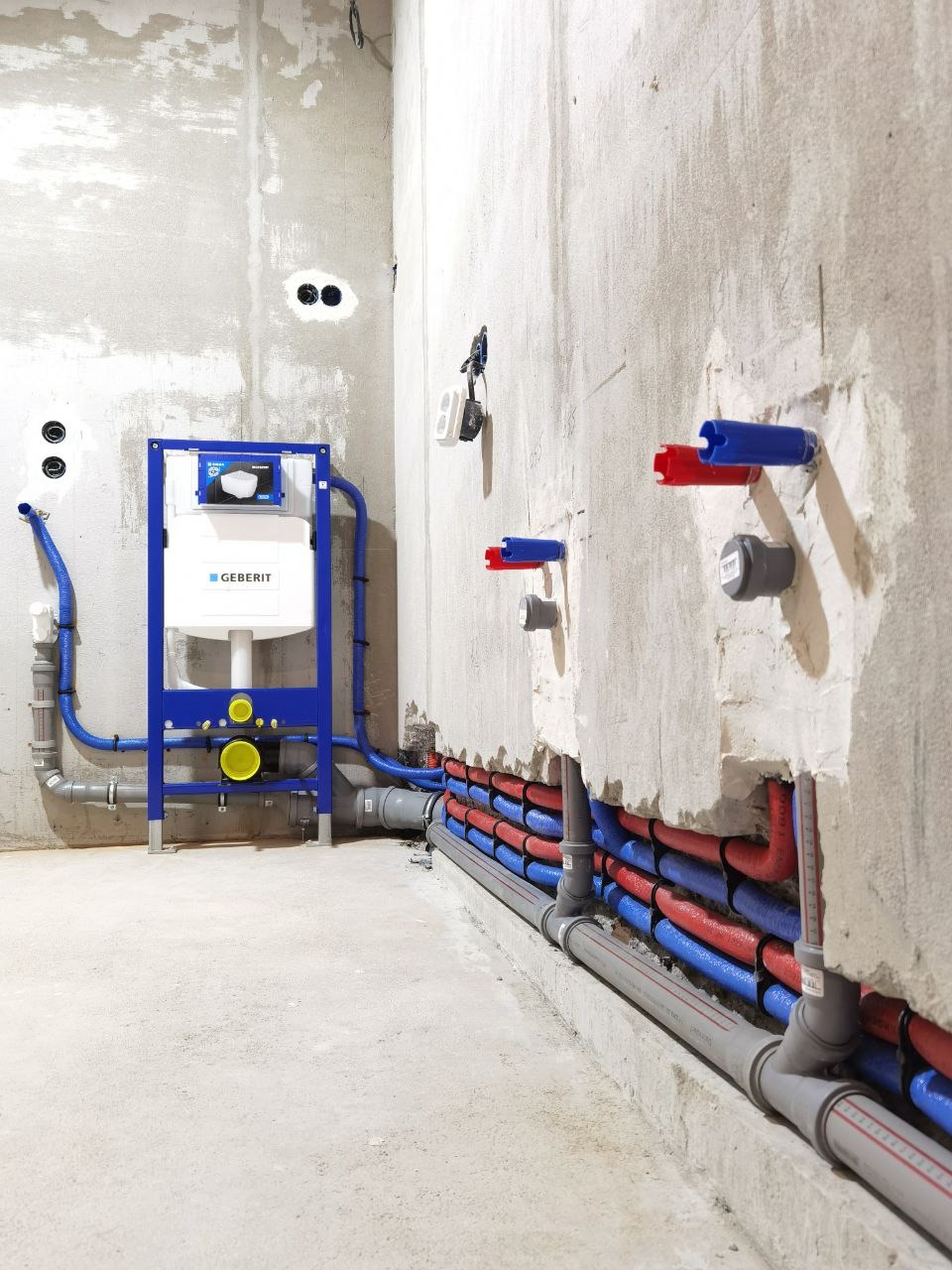 Surowy stan łazienki z widoczną instalacją hydrauliczną: stelaż Geberit, rury w izolacji czerwonej i niebieskiej, szare rury odpływowe. Ściany w stanie deweloperskim.