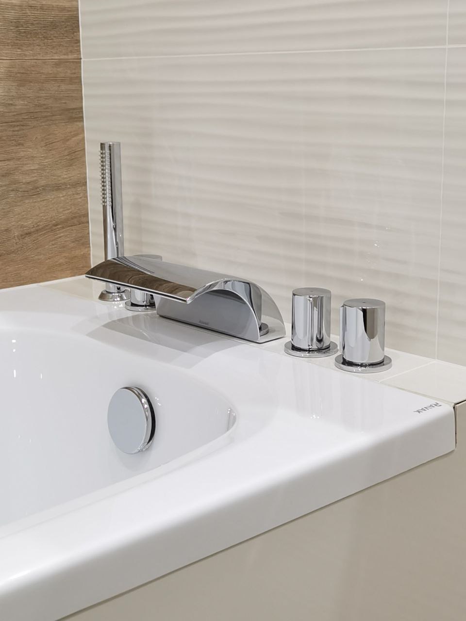 Bateria wannowa chromowana, czterootworowa, z minimalistycznymi pokrętłami i słuchawką prysznicową, na tle płytek w odcieniach beżu i brązu.