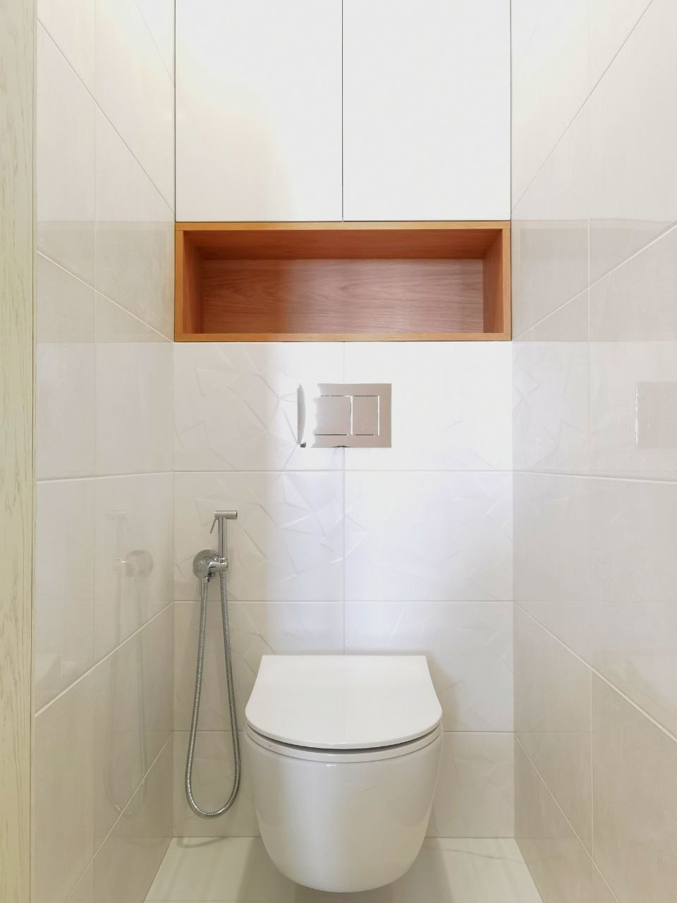 Nowoczesna toaleta podwieszana z bidetką i białymi płytkami z subtelnym wzorem, uzupełniona drewnianą półką i białymi szafkami. Minimalistyczny design, czystość i elegancja.